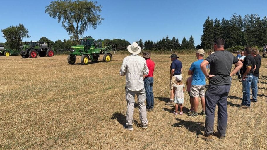 Spectators, including a small child, stand in a harvested field watching two self-propelled sprayers, a John Deere and a Fendt, drive past. Photo: John Greig
