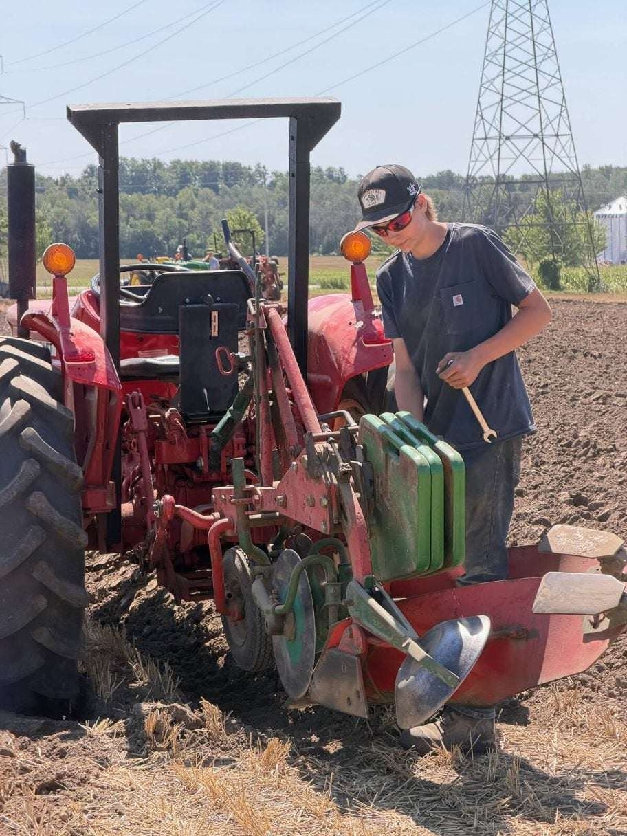 Aiden Hickey, in a ball cap and sunglasses, holds a wrench as he adjusts the plow hitched to his red tractor. Photo: John Greig