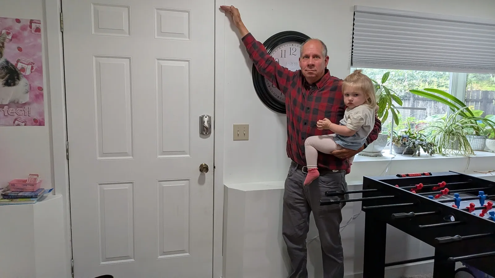 A man in a plaid shirt holding a child points to a mark on the wall of his home to show how high floodwaters rose