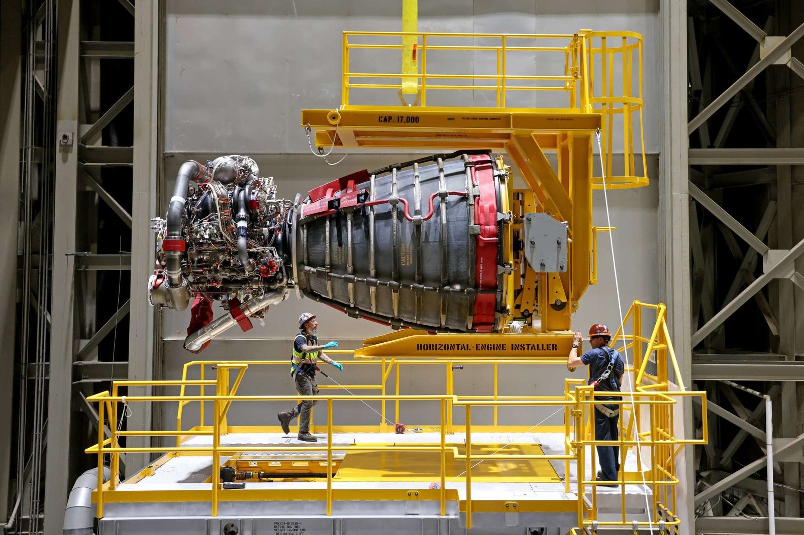 Two technicians at NASA’s Michoud Assembly Facility in New Orleans stand around one of the four RS-25 engines for the agency’s SLS (Space Launch System) rocket on Sept. 8, 2023. The RS-25 engine looks like a bell-shaped nozzle attached to a network of pipes. The engine is being lifted by the horizontal engine installer so it appears to be lying on its side. The RS-25 engine is about the size of a large pickup truck. The technicians are wearing hard hats and safety harnesses.