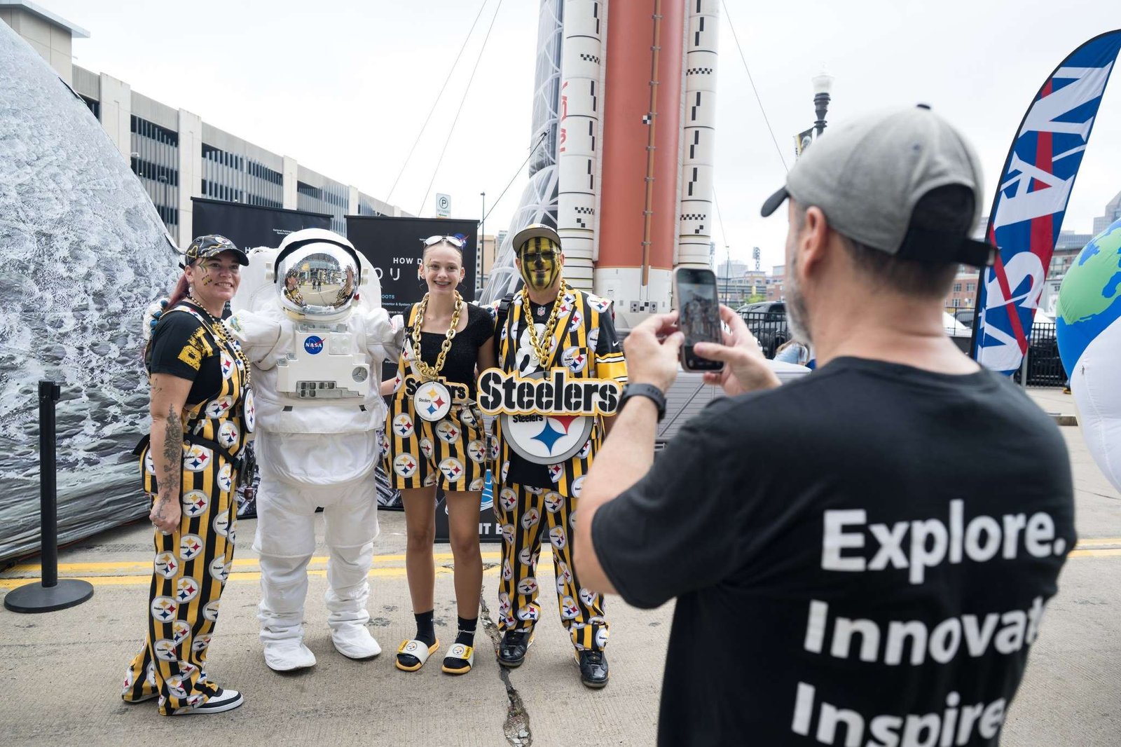 Three Pittsburgh Steelers fans, dressed in black and yellow clothes with the Steelers' logo on it, pose with an astronaut in a spacesuit. The reflective visor is closed. In the foreground, a man takes the group's picture with his phone.