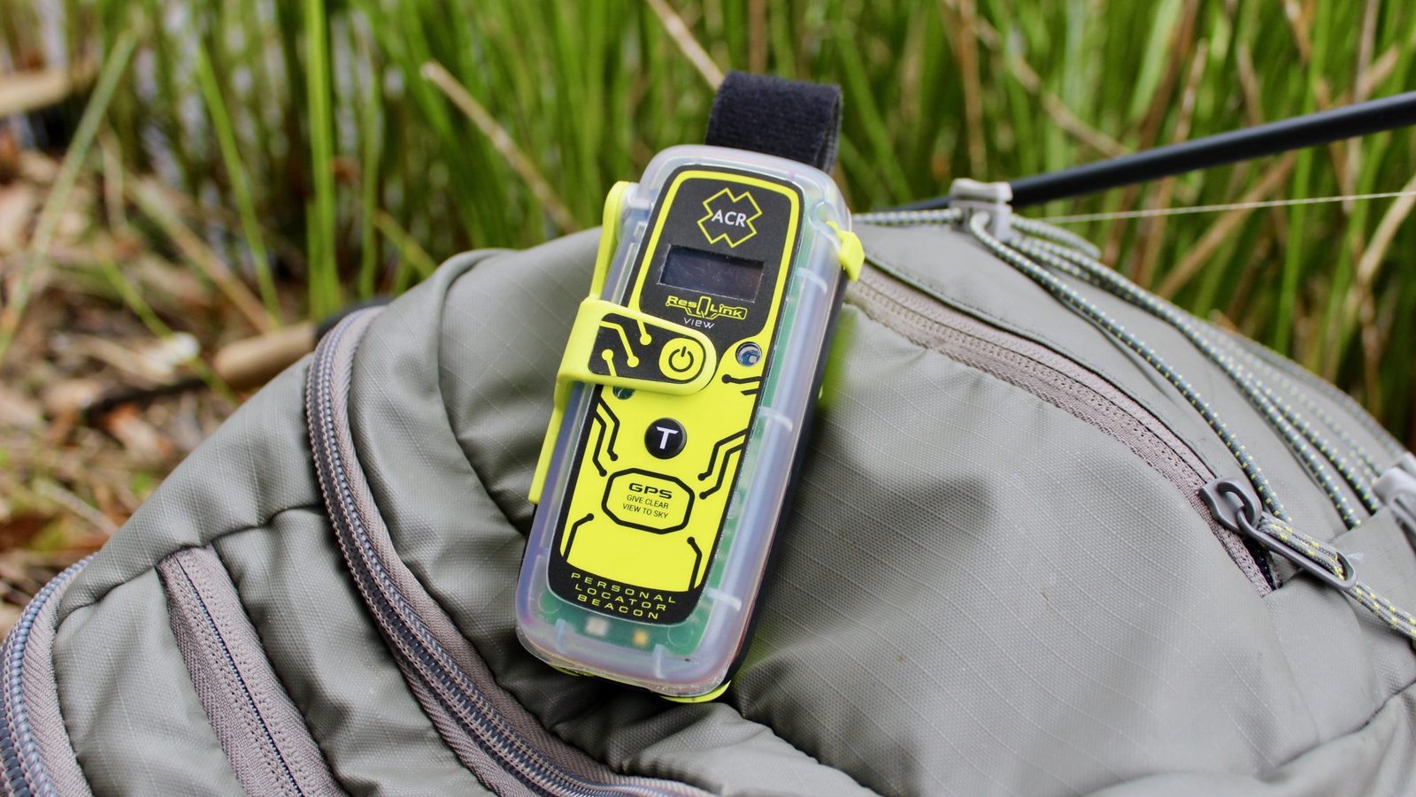 A close up picture of a green ACR PLB atop a bag in a forest setting