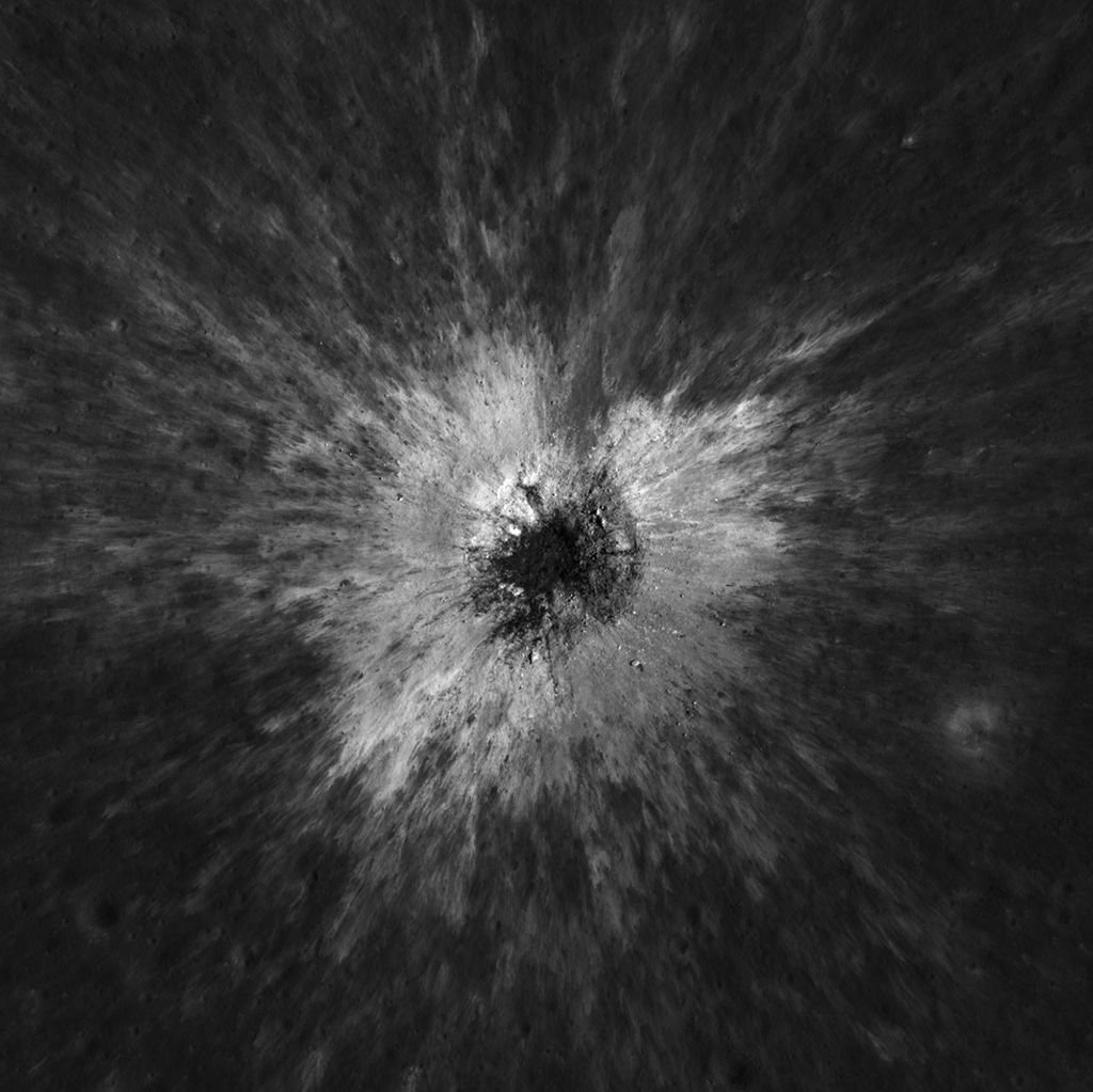 Square black-and-white image of a dark, uneven surface. At the center is a dark, roughly circular depression surrounded by a bright irregular area, with long streaks of lighter material radiating outward in all directions.