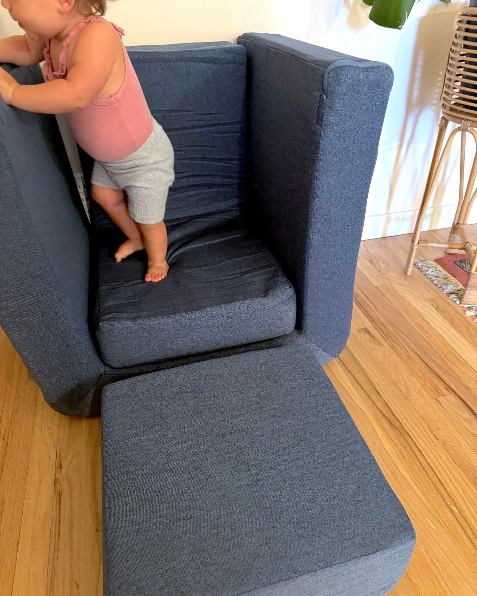 A toddler in a pink top and gray shorts stands on a dark blue foam couch with one cushion pulled out onto a wooden floor.