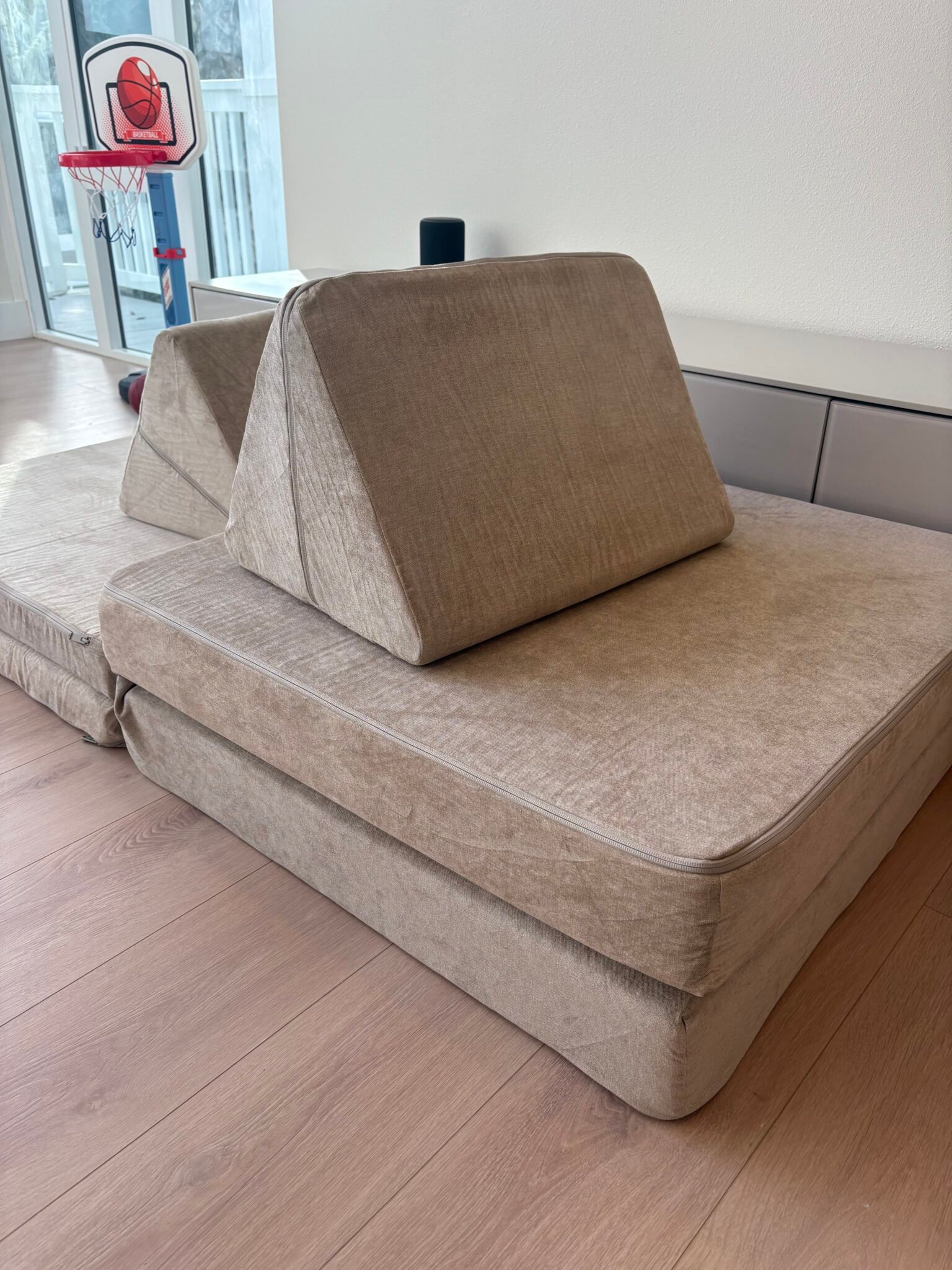 A beige modular foam couch with triangular back supports is set on a wooden floor near a small indoor basketball hoop by a window.
