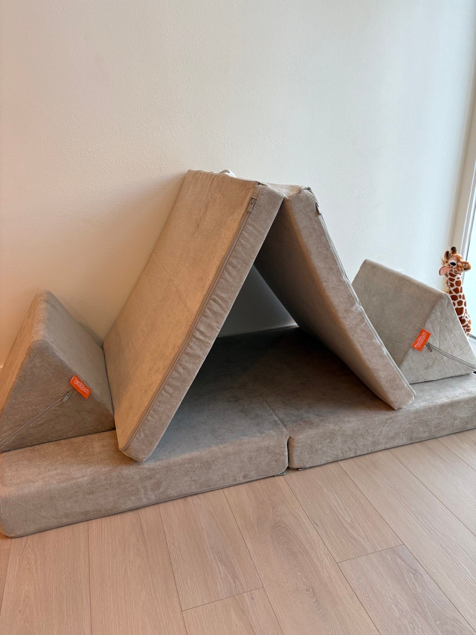 A foldable foam play couch is arranged as a tent with two triangular cushions on the sides. A toy giraffe is visible in the background near the wall.