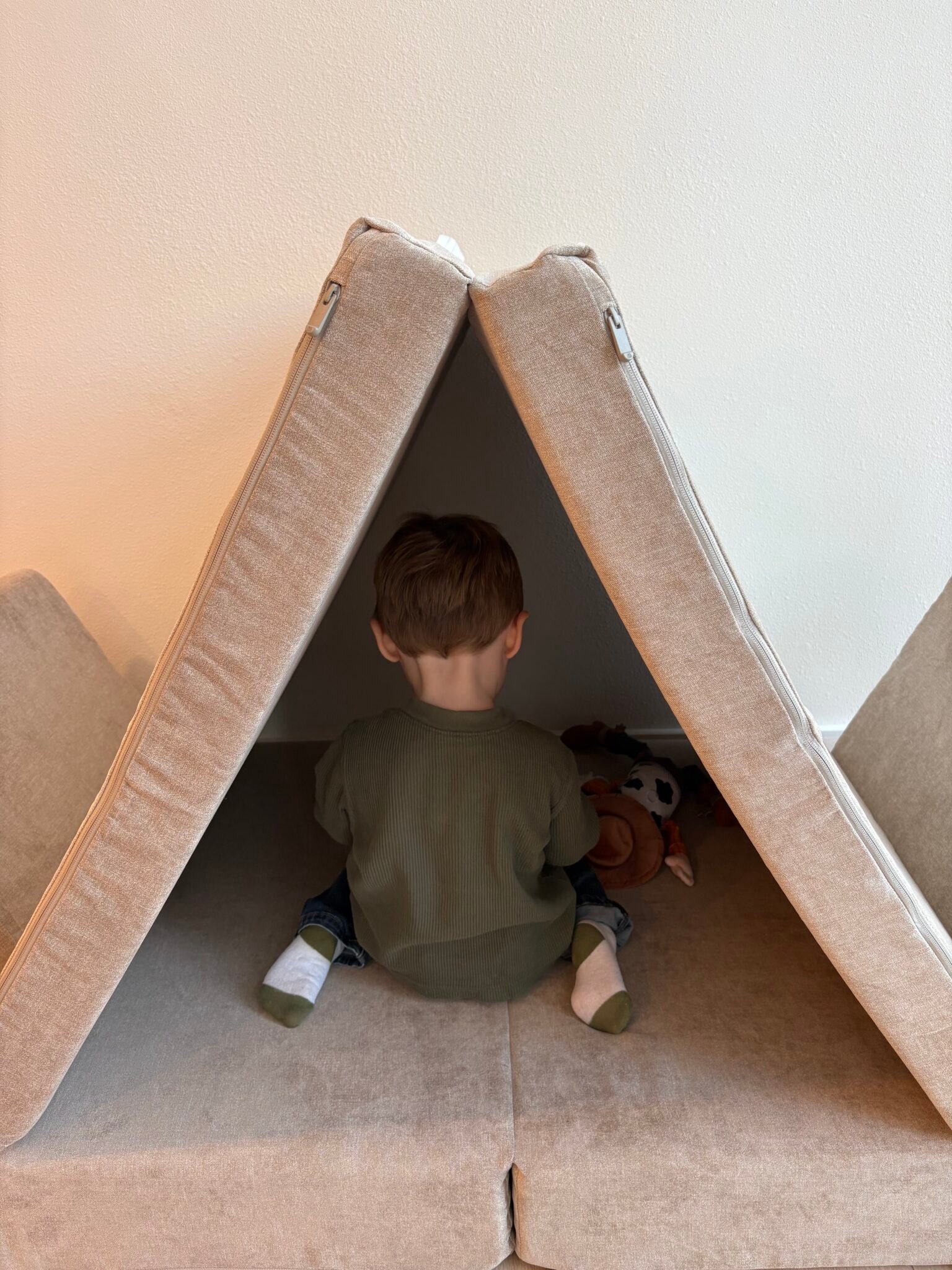 A young child sits with their back to the camera inside a makeshift tent made of beige couch cushions, playing with stuffed animals.