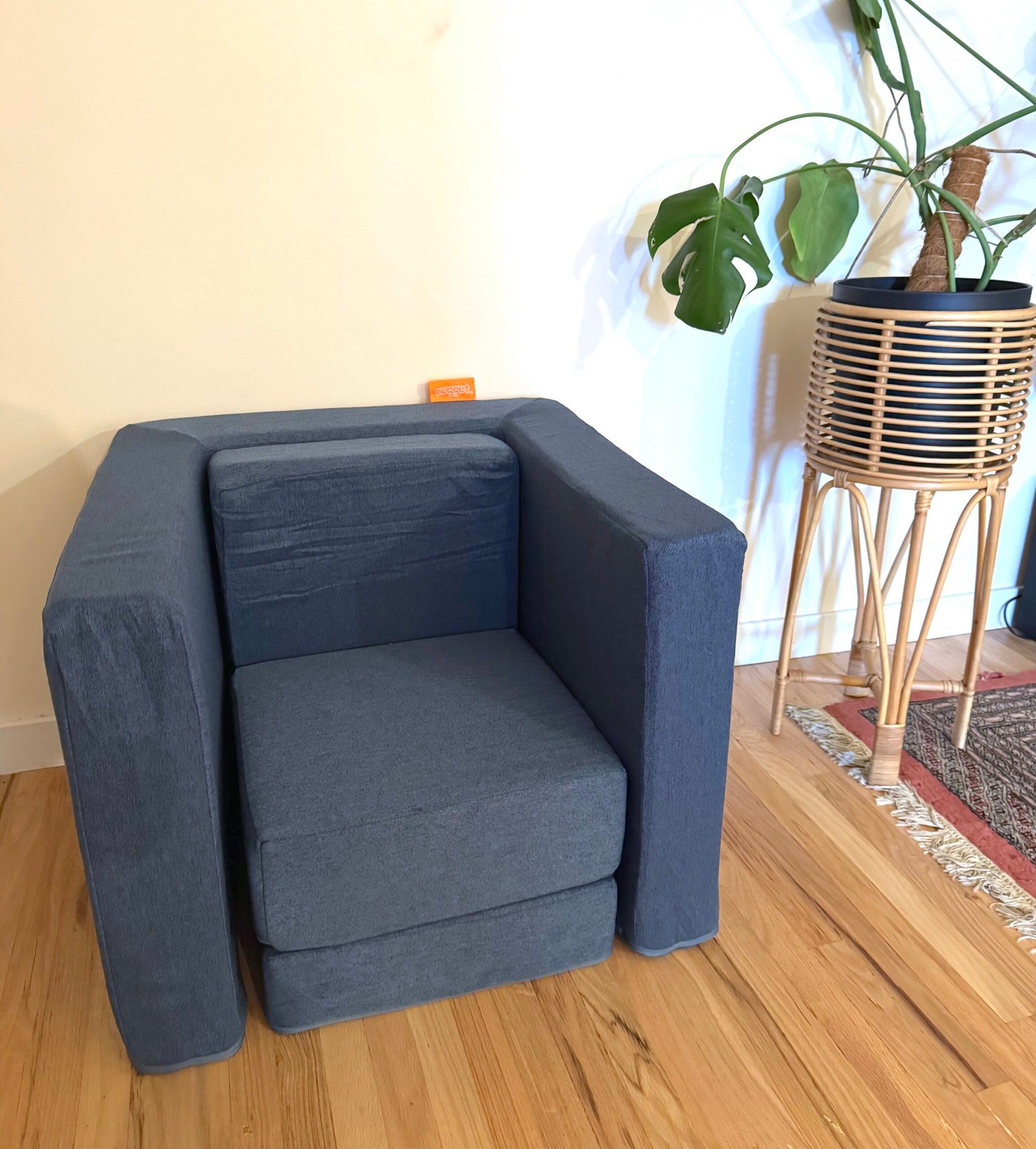 A dark gray armchair sits on a wooden floor near a potted plant in a wicker stand, against a light-colored wall.