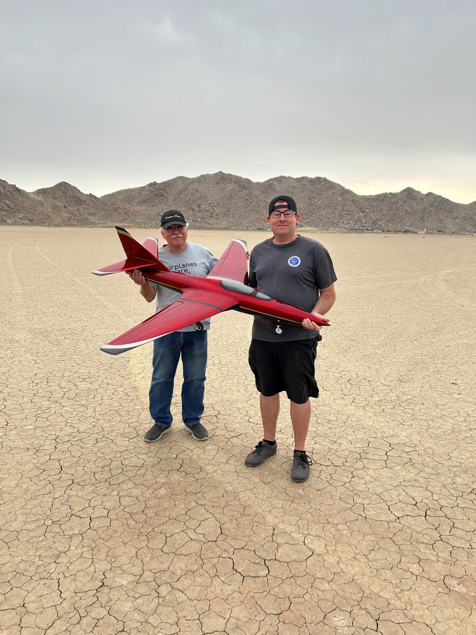 Two men hold a large radio-controlled aircraft. The aircraft is red with white and black stripes.