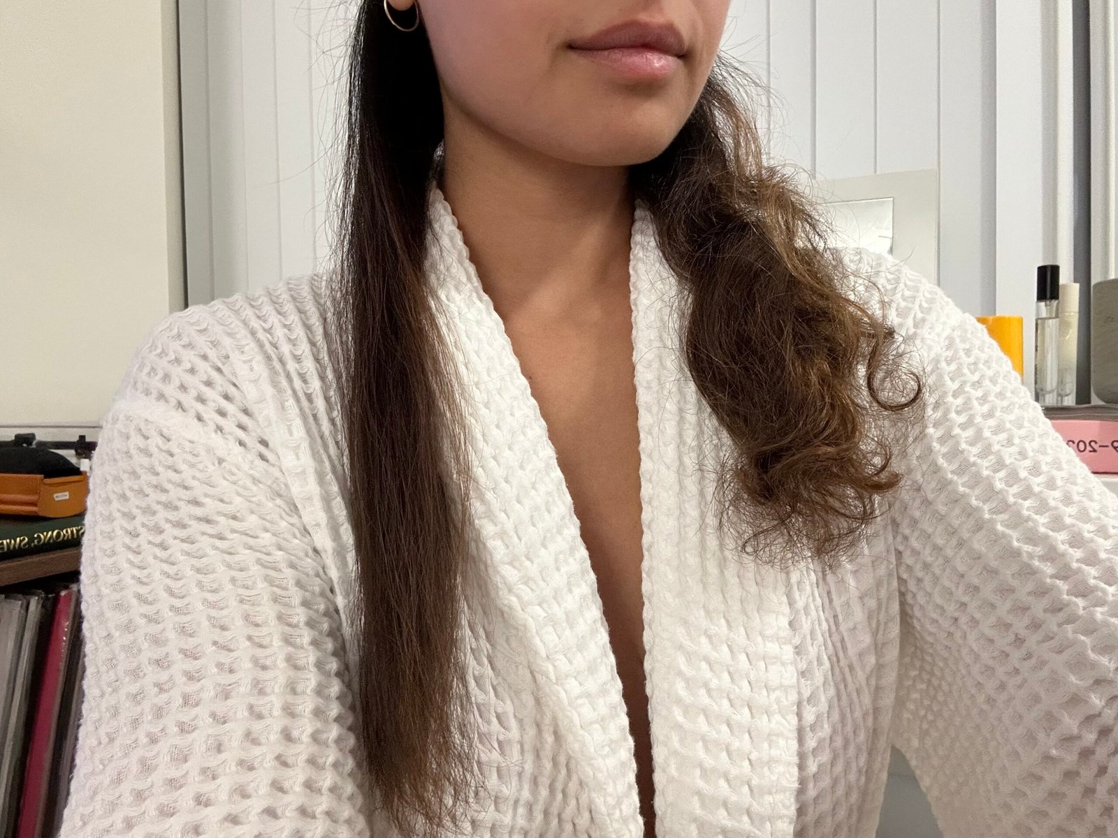 A person in a white robe shows their hair, with one half straight and the other half curly, against a background of shelves and books.