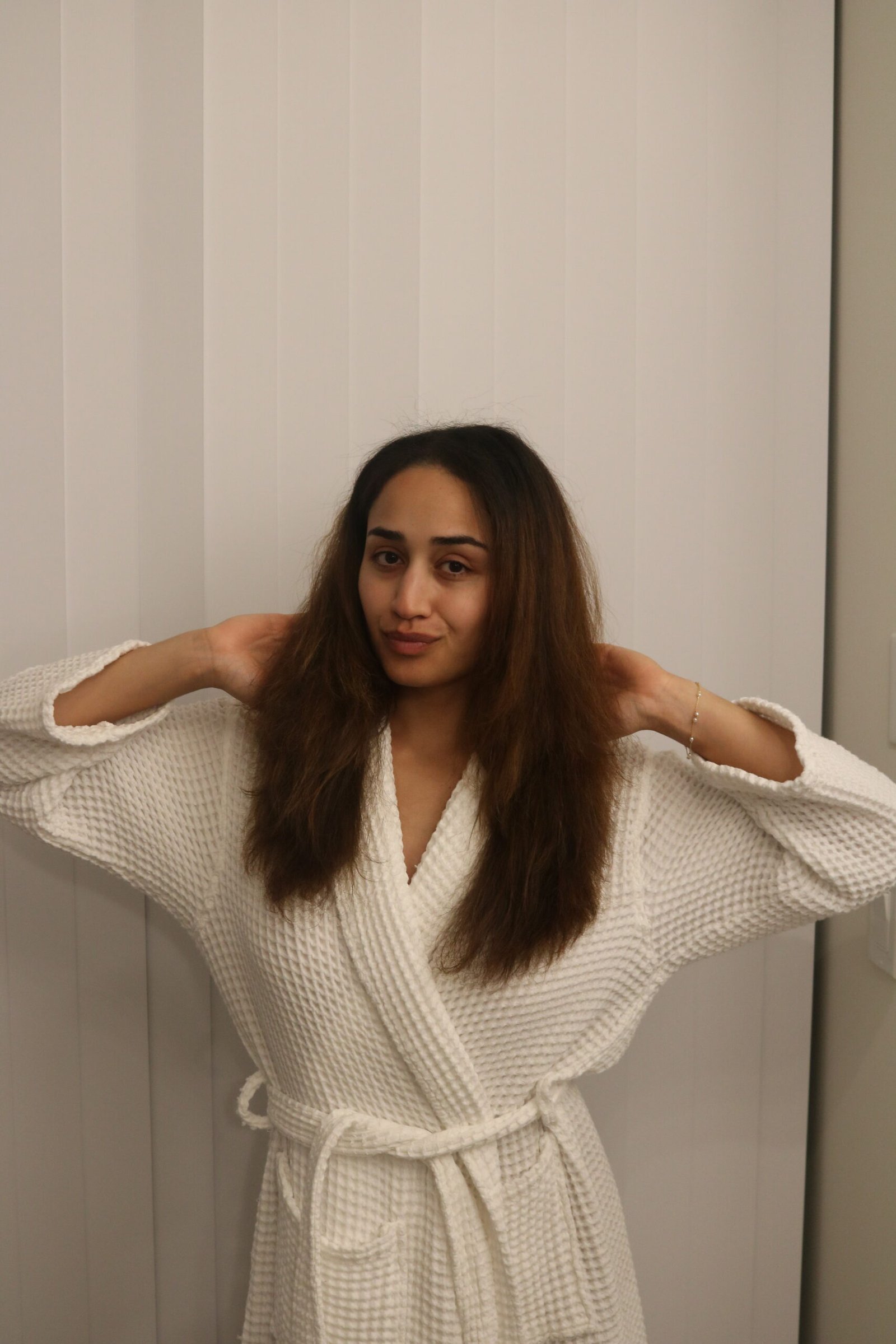 A person wearing a white bathrobe stands indoors, facing the camera with their hands raised to touch their hair, in front of vertical white blinds.