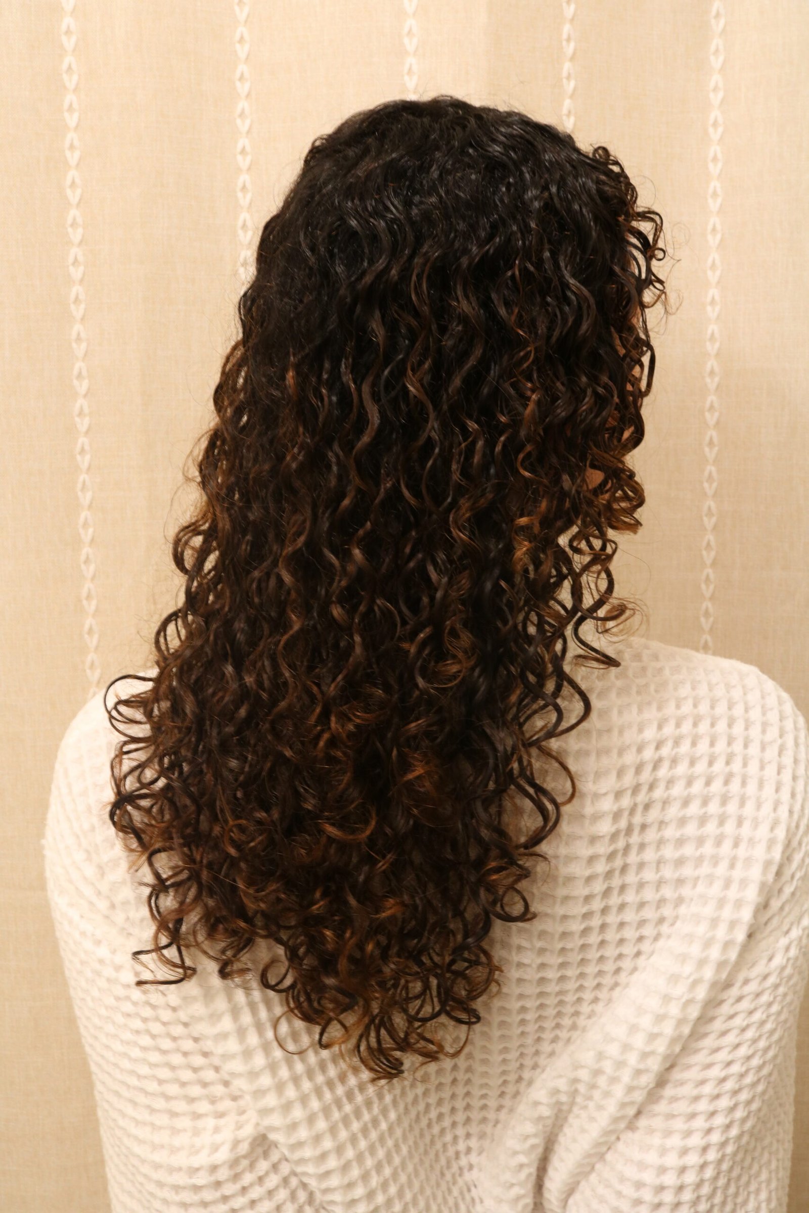 A person with long, dark, tightly curled hair is shown from the back, wearing a textured white top against a beige patterned background.