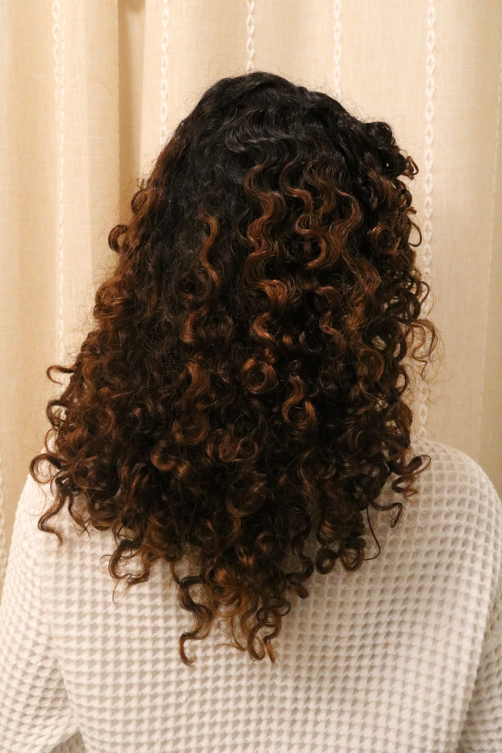 Person with shoulder-length, dark brown and light brown curly hair facing away, wearing a white textured sweater in front of a beige curtain.