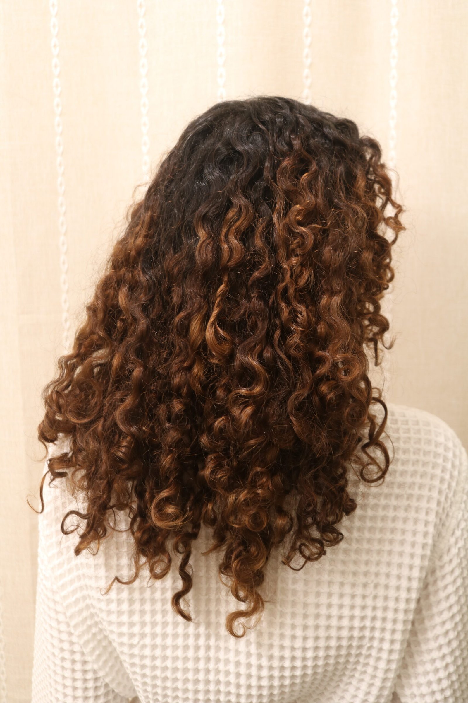 Person with shoulder-length, dark brown curly hair featuring lighter brown highlights, wearing a textured white top, shown from behind against a beige background.