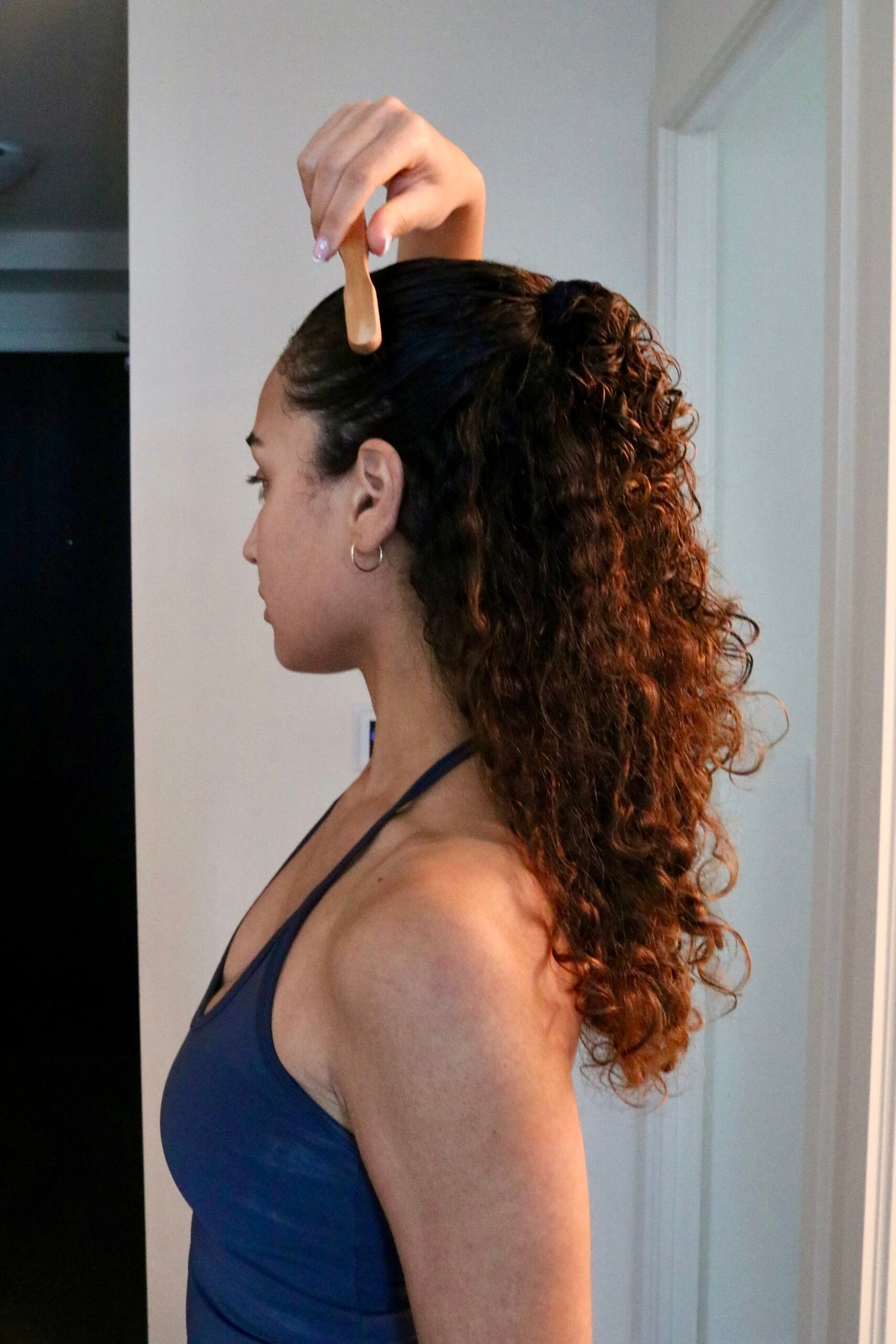 Woman with curly hair styles her hair using a comb, holding it at the top of her head, standing in a hallway and wearing a blue tank top.
