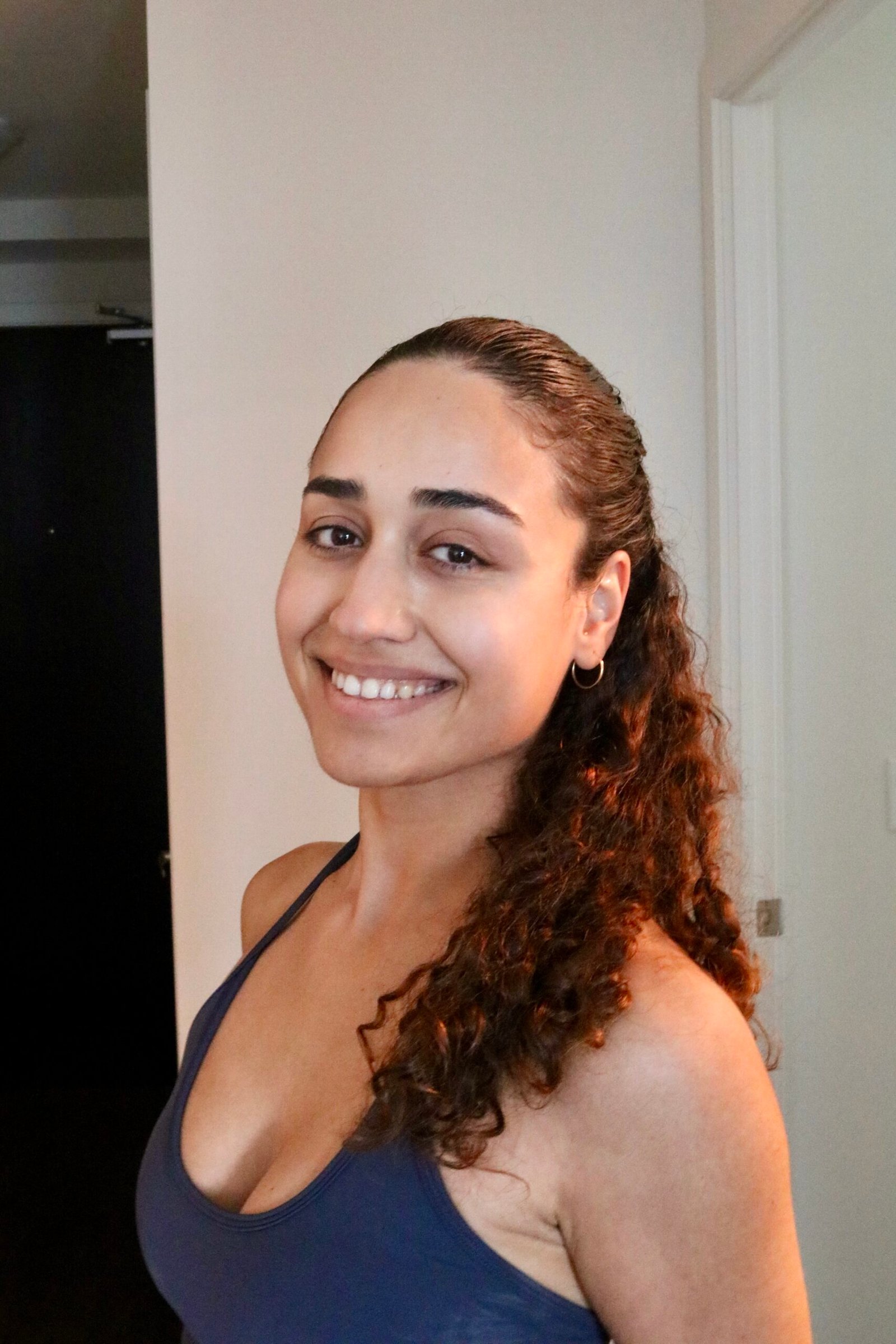 Woman with curly hair wearing a navy tank top smiles while standing indoors near a white wall and doorframe.