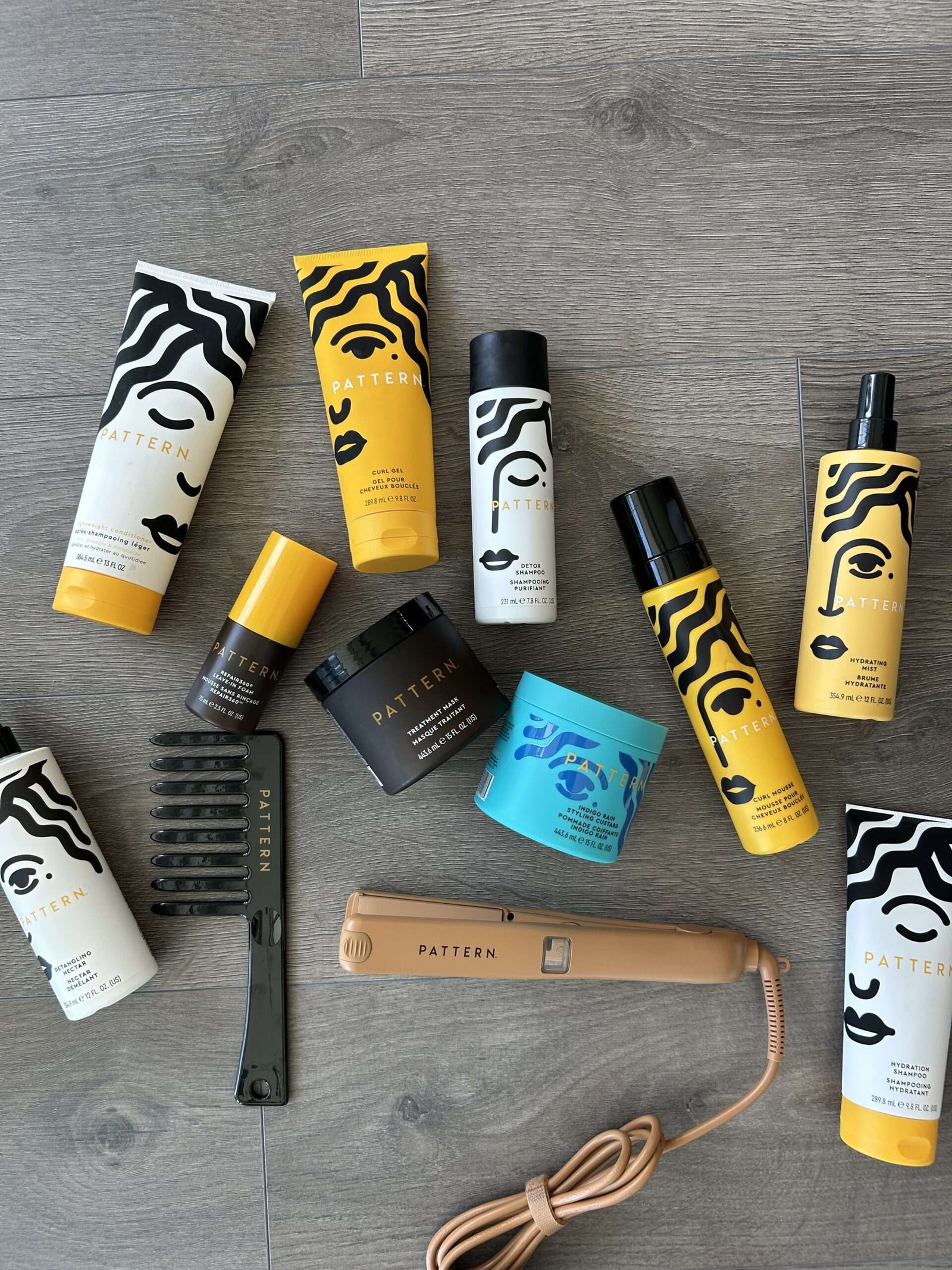 A collection of Pattern and Ampro hair products, a wide-tooth comb, a hair pick, and a tan Pattern flat iron arranged on a wooden floor.