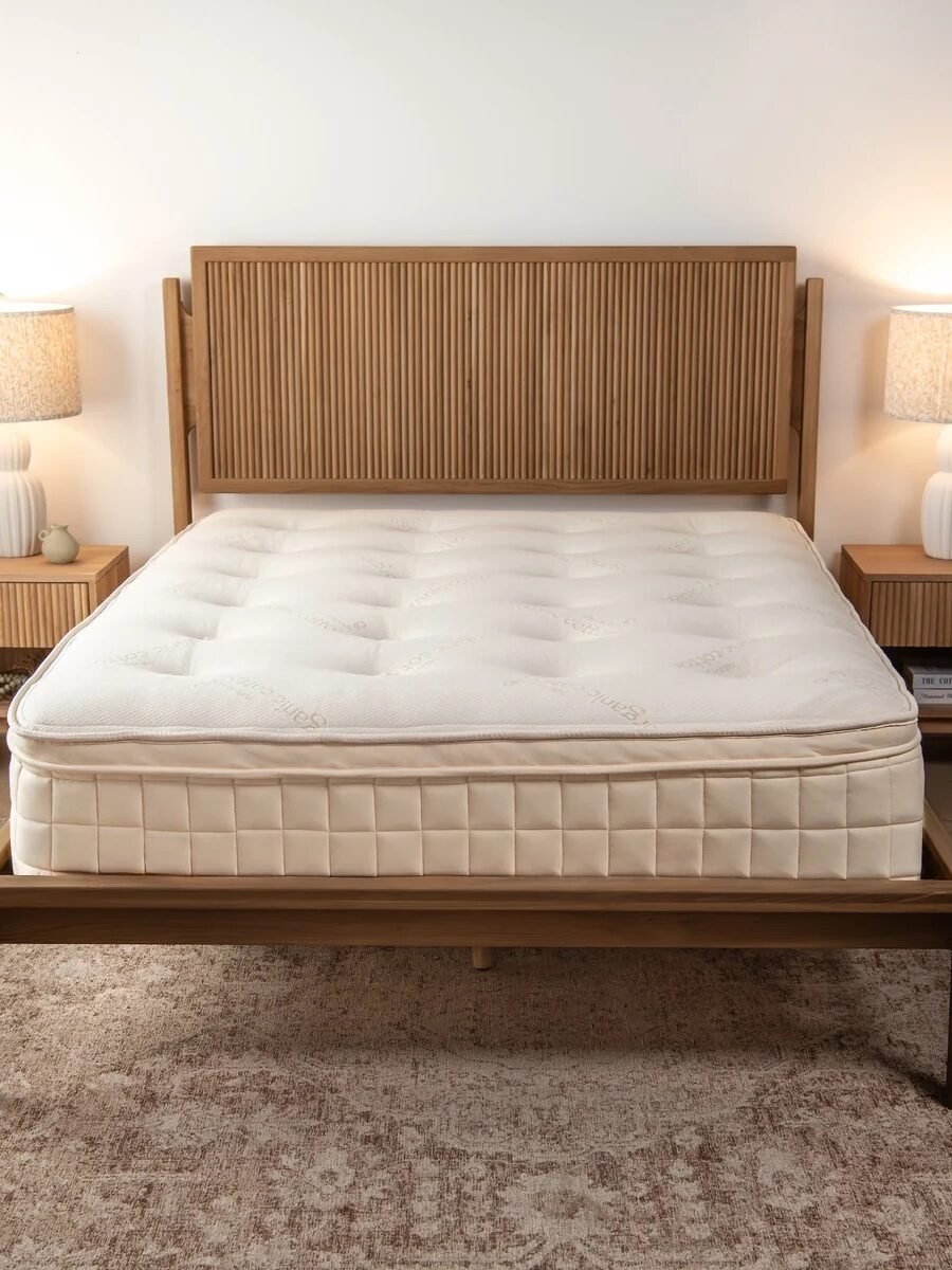 A neatly made bed with a wood frame sits between two matching nightstands with lamps, placed on a beige rug in a simple, well-lit bedroom.