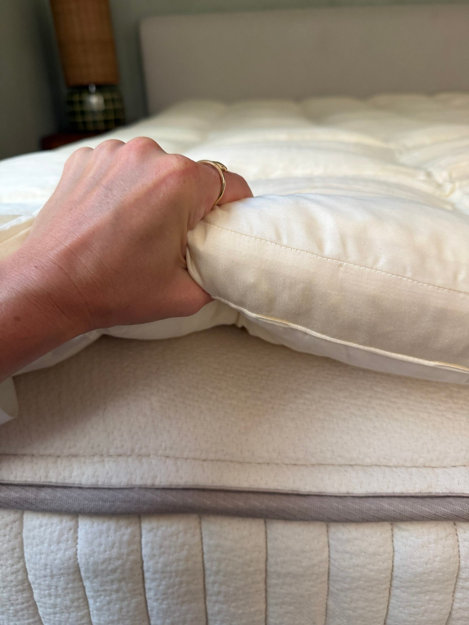 A hand gripping the edge of a thick, beige mattress topper placed on top of a mattress.
