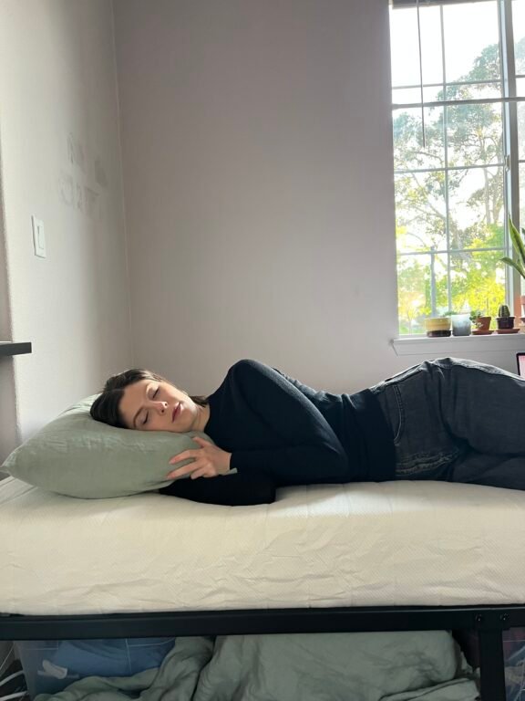 Person lying on their side on a bed with a green pillow, eyes closed, wearing a black top and gray jeans, next to a window with sunlight and potted plants visible outside.