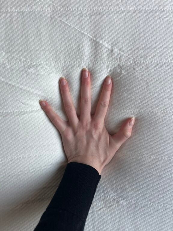 A hand with fingers spread is pressing down on a textured white surface, possibly a mattress or cushion.