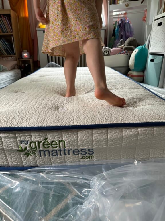 A child stands barefoot on a Green Mattress in a bedroom, with toys and shelves visible in the background.