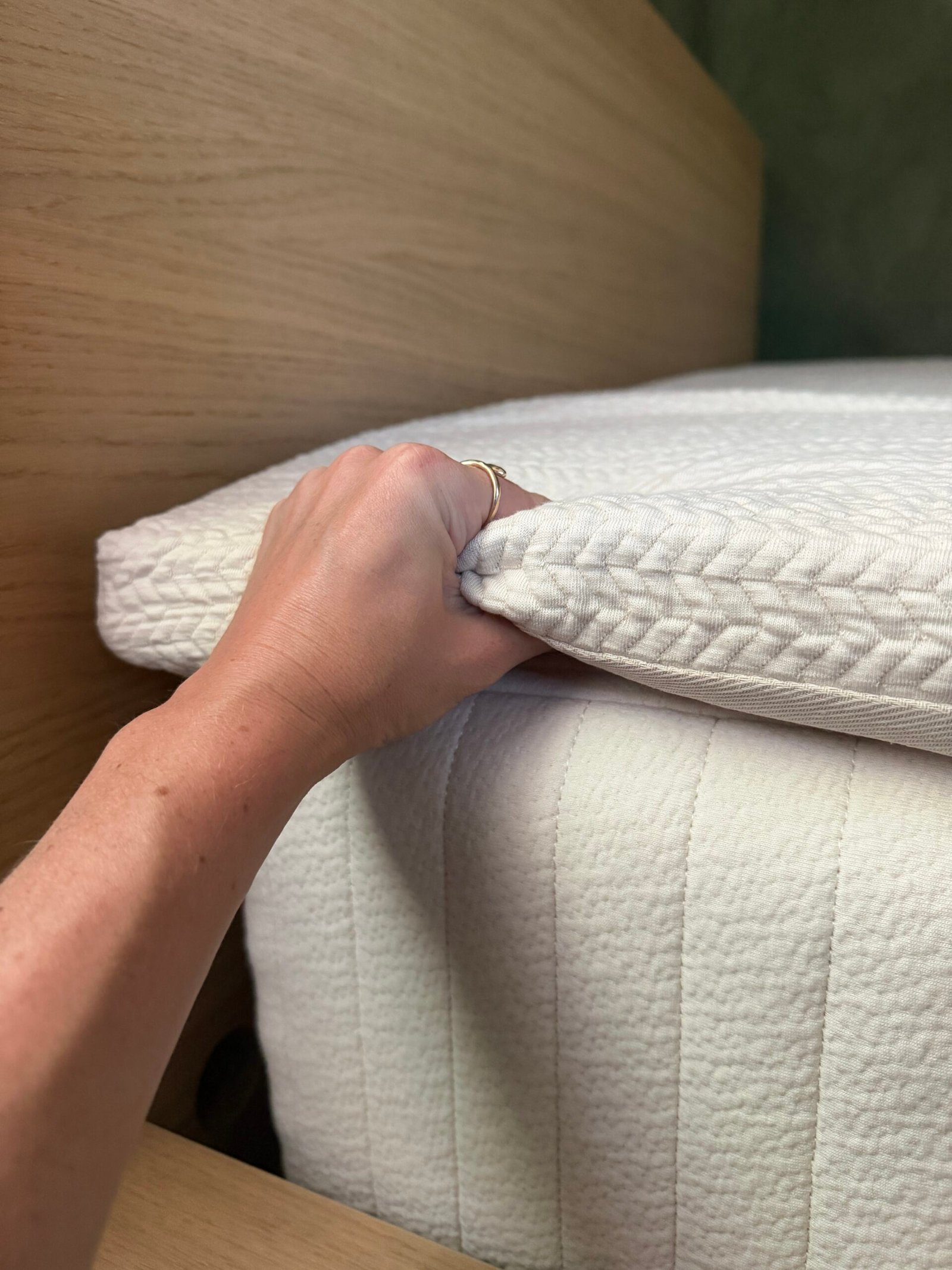 A hand lifts the corner of a white mattress topper, revealing its texture and thickness on top of a mattress.