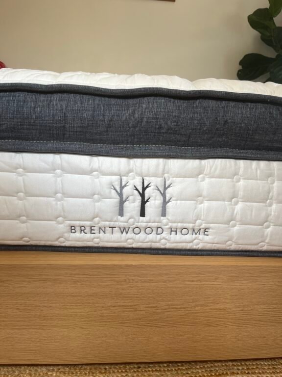 Side view of a Brentwood Home mattress on a wooden bed frame, showing quilted fabric and the brand’s tree logo embroidered in gray.