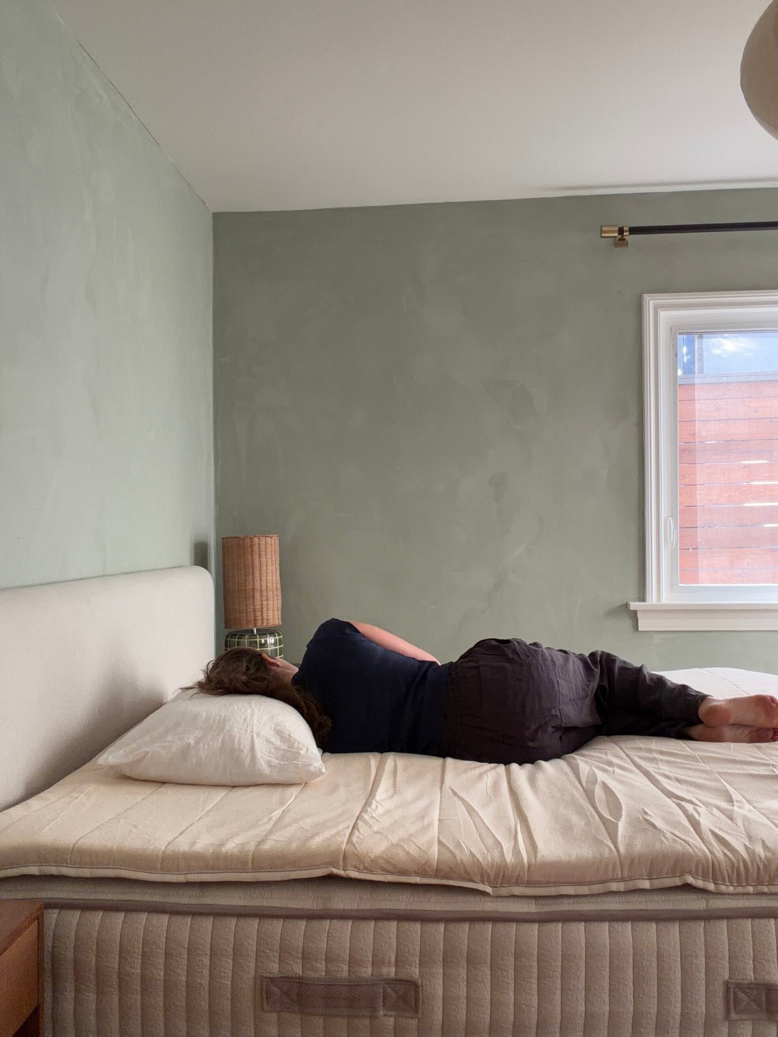 Person lying on their side on an unmade bed in a sparsely furnished bedroom with a light green wall and window.