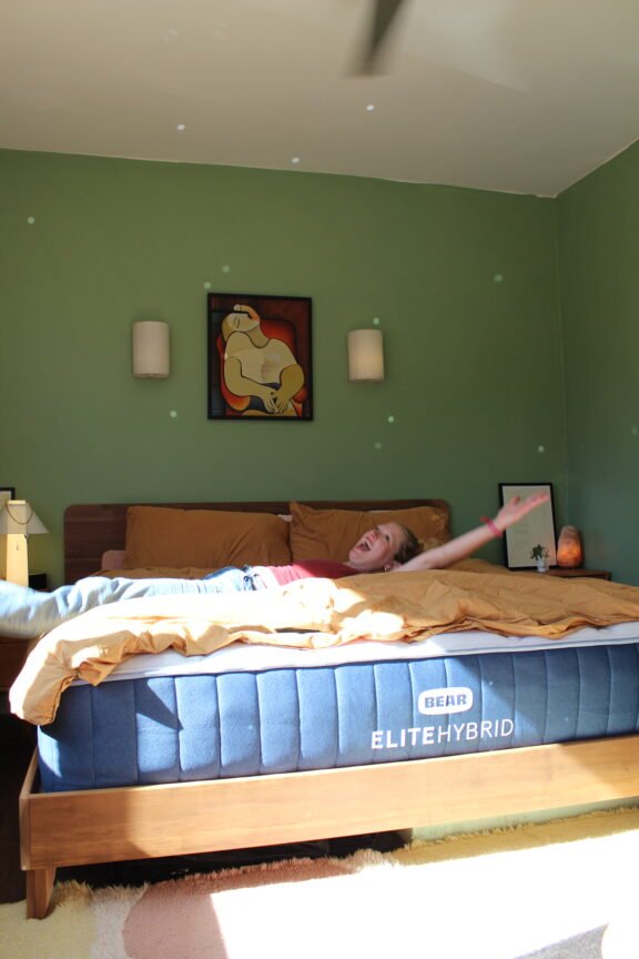 Person lying on a Bear Elite Hybrid bed in a sunlit bedroom with green walls, mustard bedding, and art above the headboard.