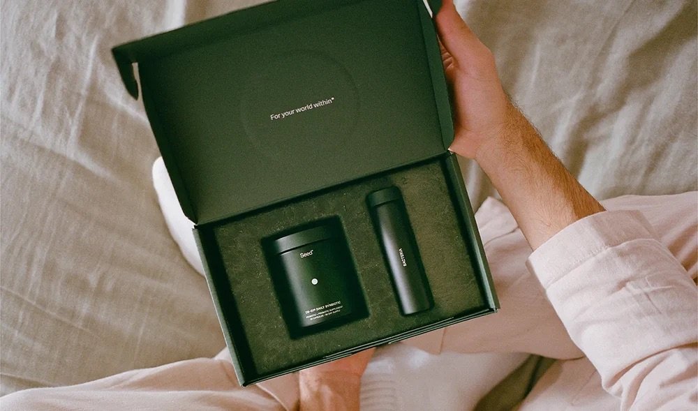 A person in light clothing holds an open box containing a black candle and a black spray bottle, both labeled “Snif,” on a beige fabric surface—perfect for discovering new favorites during the Labor Day sale.