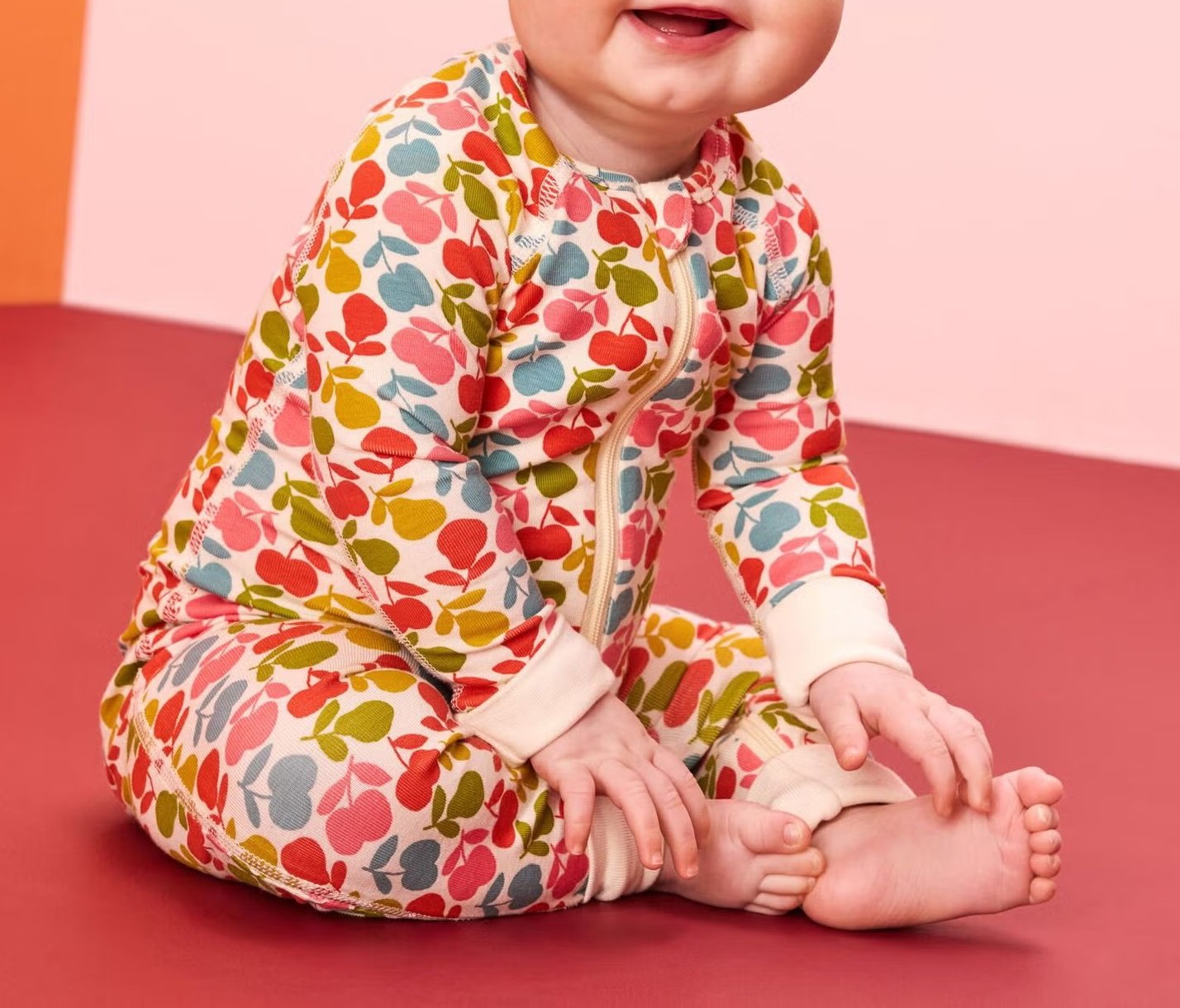 A baby sits on a red surface wearing a long-sleeve, zip-up onesie with a colorful leaf pattern in red, yellow, green, and pink.