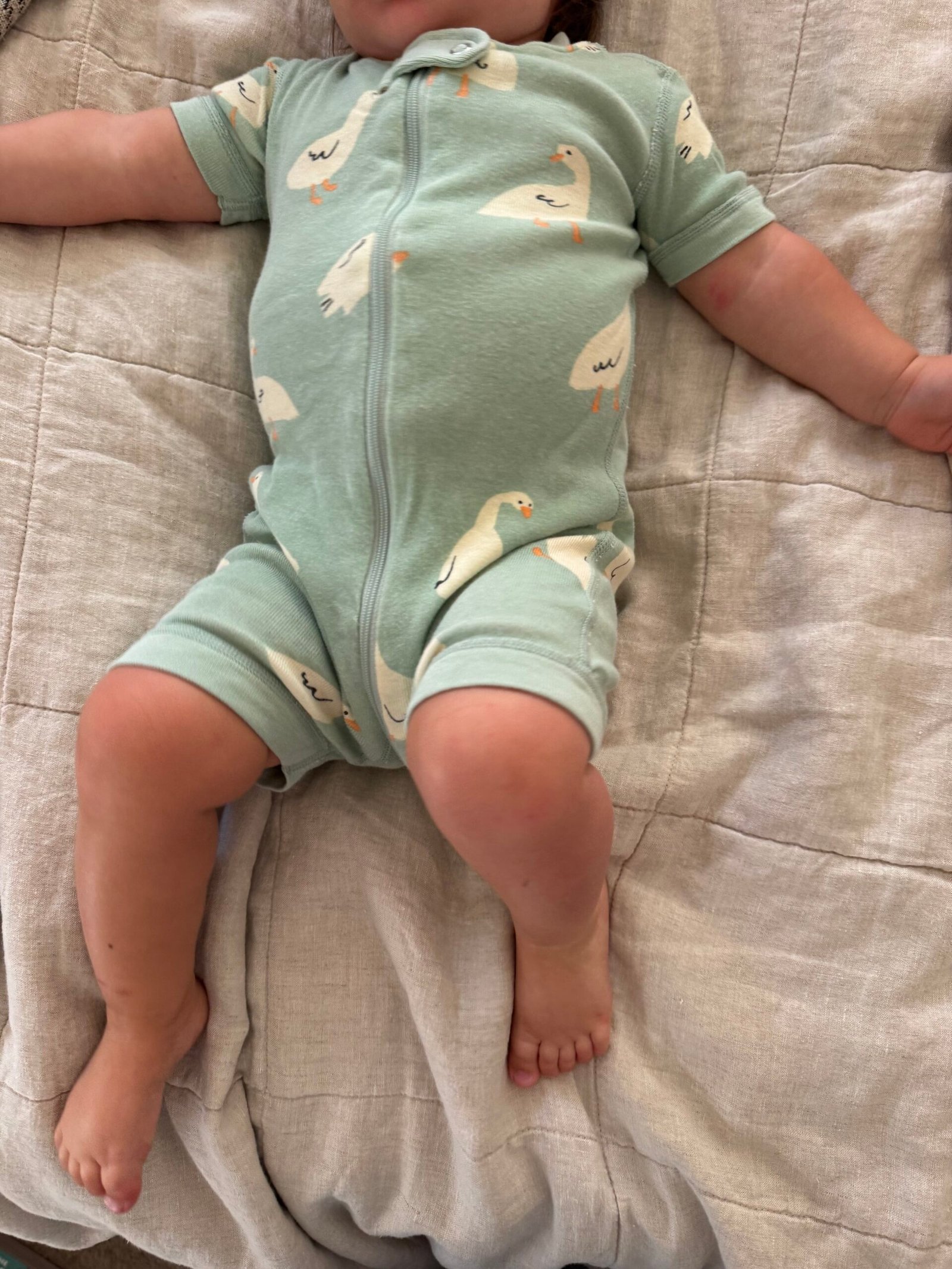 Baby lying on a beige blanket, wearing a light green romper with a duck print and short sleeves, arms outstretched and legs slightly apart.