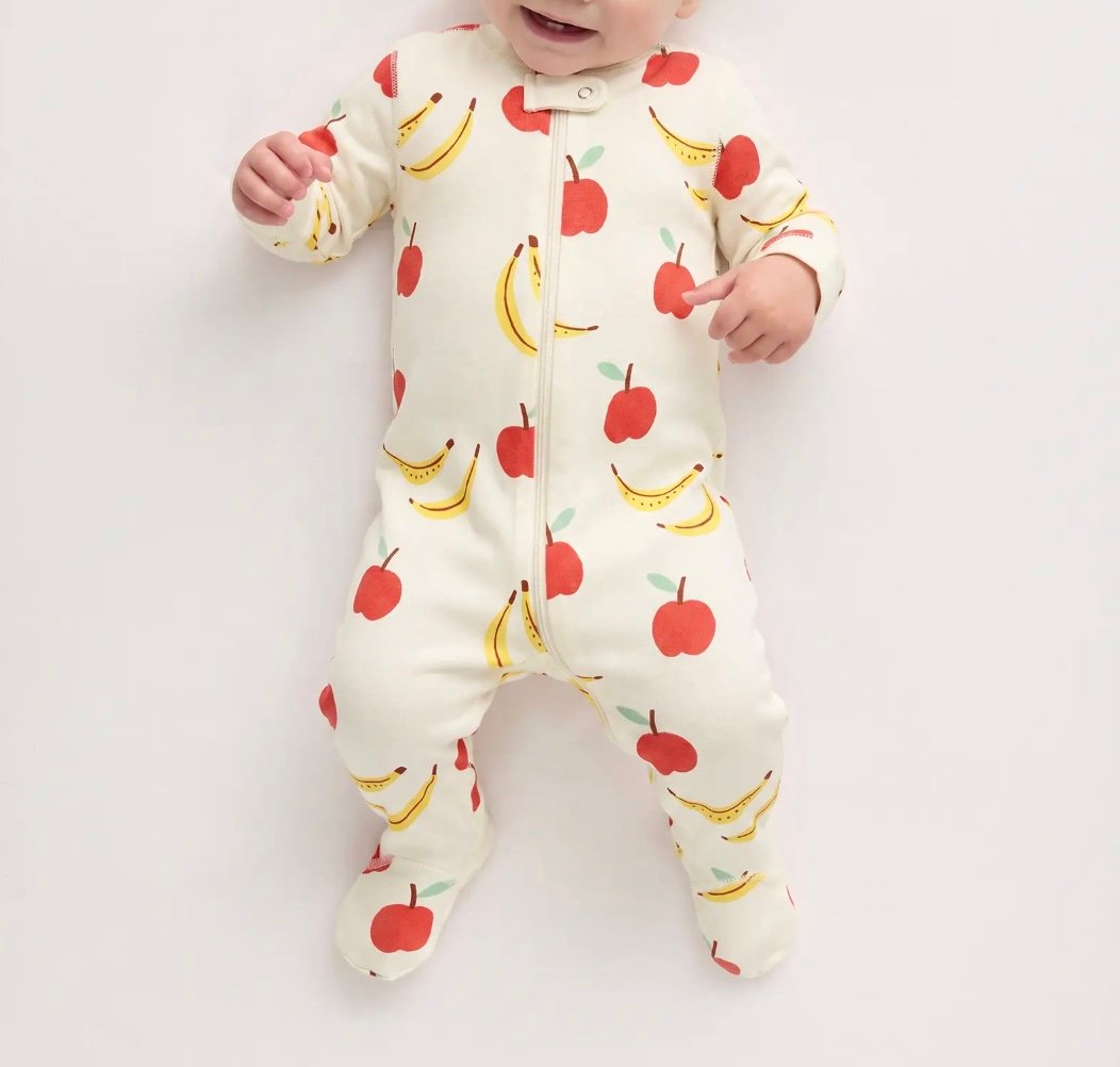 Baby wearing a cream-colored footed onesie with red apple and yellow banana print, lying on a white background.
