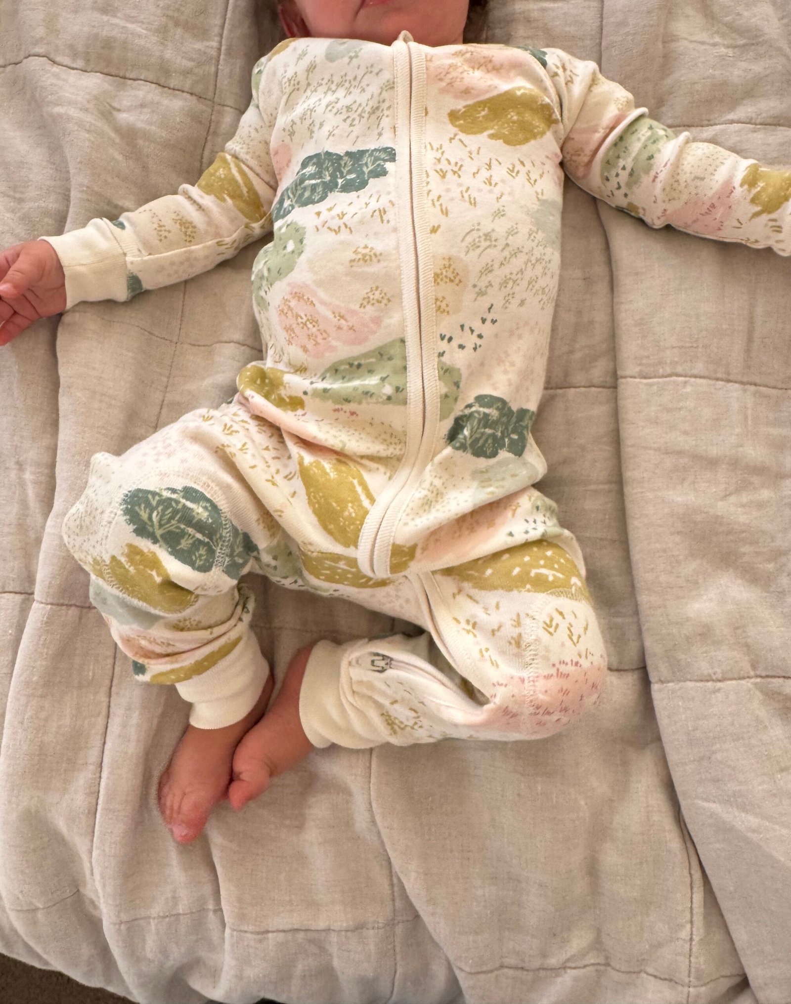 Baby in a cream-colored, patterned onesie lies on a beige quilted blanket, with arms and legs relaxed and face partially out of frame.