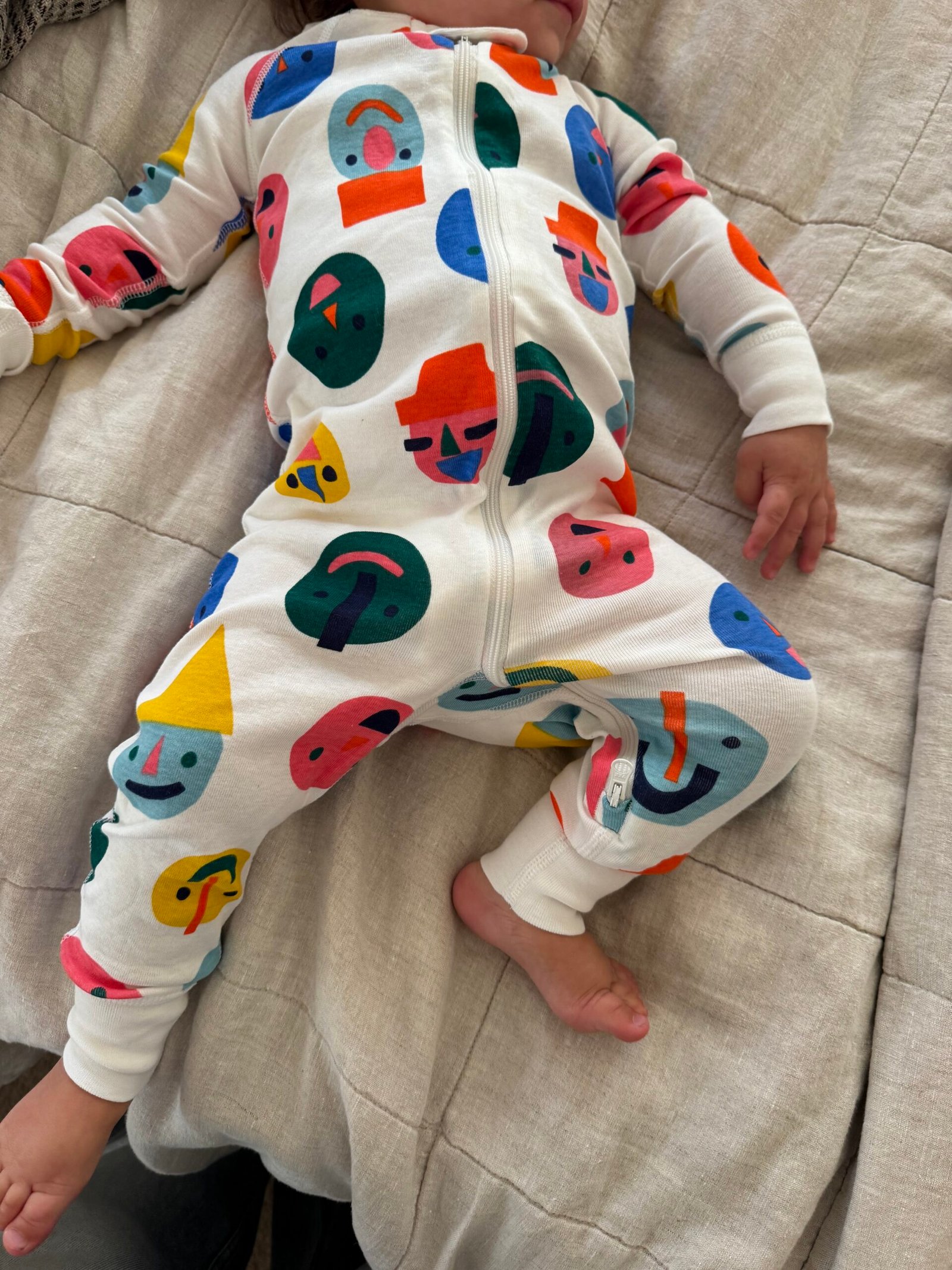 Baby lying on a beige blanket, wearing a white onesie with colorful abstract face patterns and a zipper down the front.