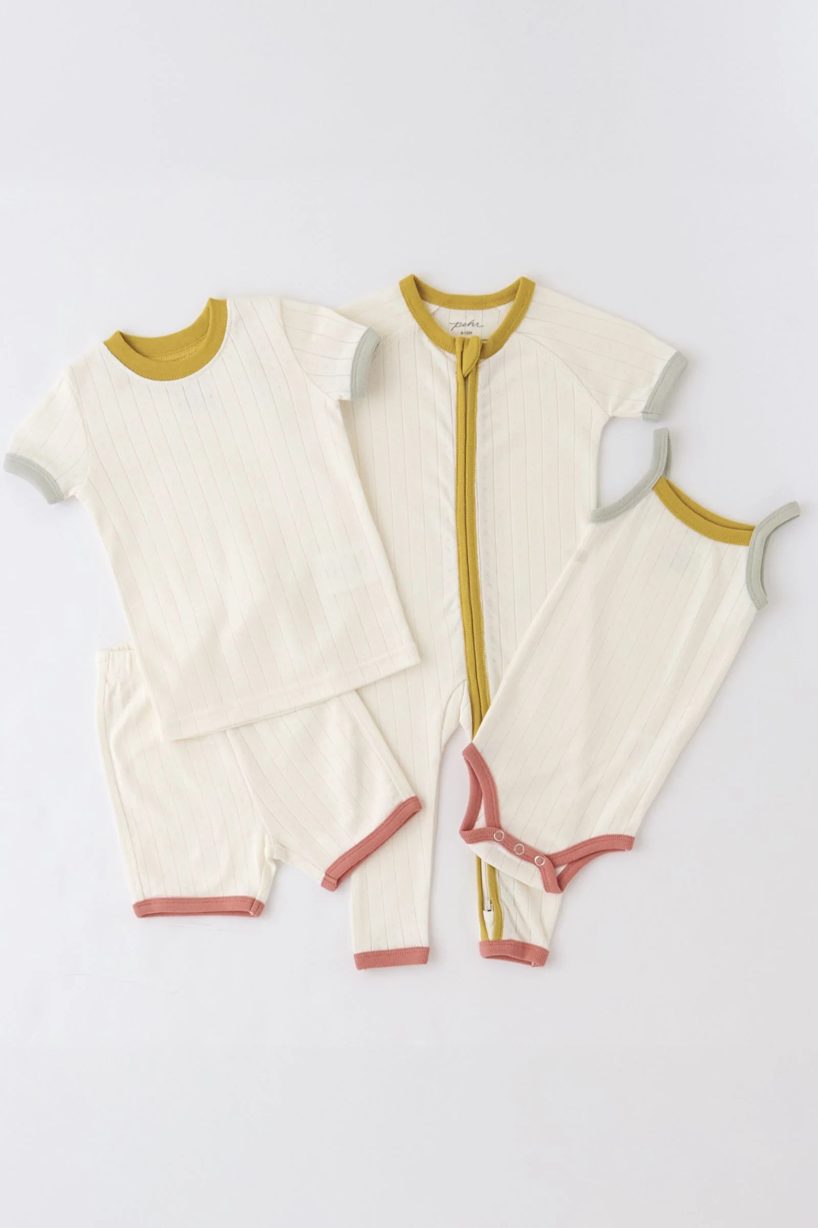 Four cream-colored baby outfits with colored trim—mustard, sage green, and pink—displayed on a white background. Styles include a romper, bodysuit, short-sleeve top, and shorts.