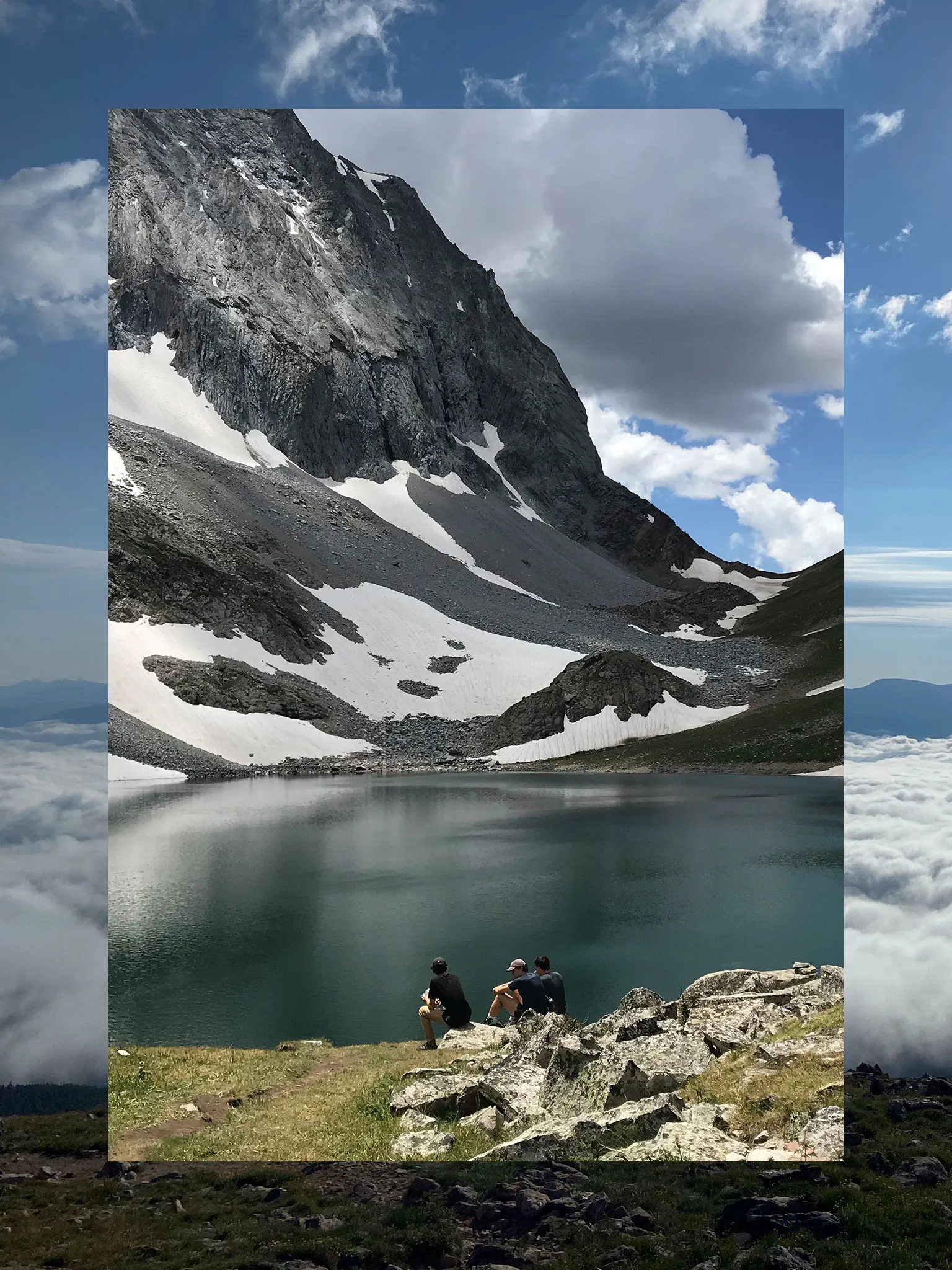A collage consisting of two vertical photos, the outside functioning a border and the inside fully visible. The outside photo is a misty mountaintop with clouds. The inner photo is of hikers sitting beside a lake.