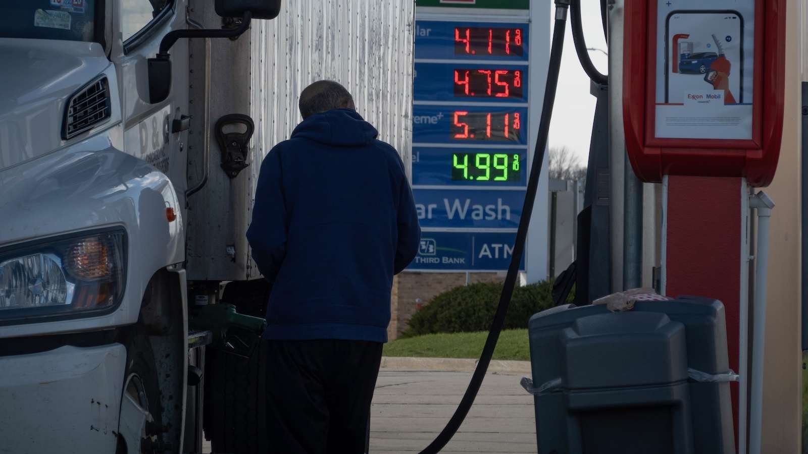 A truck driver fills his big-rig with diesel fuel. A sign in the background indicates the price is four dollars and ninety-nine cents a gallon.