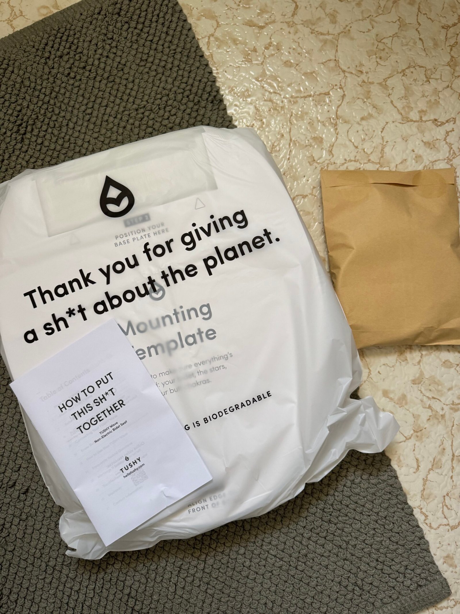 Biodegradable package on a bath mat with a paper guide titled “HOW TO PUT THIS SH*T TOGETHER” and a brown envelope, all on a tiled floor.