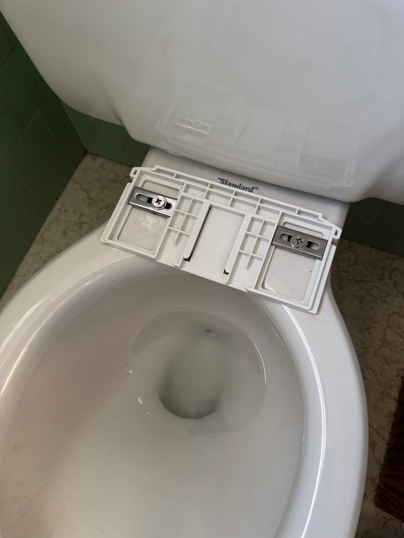 A toilet with its seat removed, exposing the mounting bracket and screws on the rim, in a bathroom with green tile walls and a beige patterned floor.