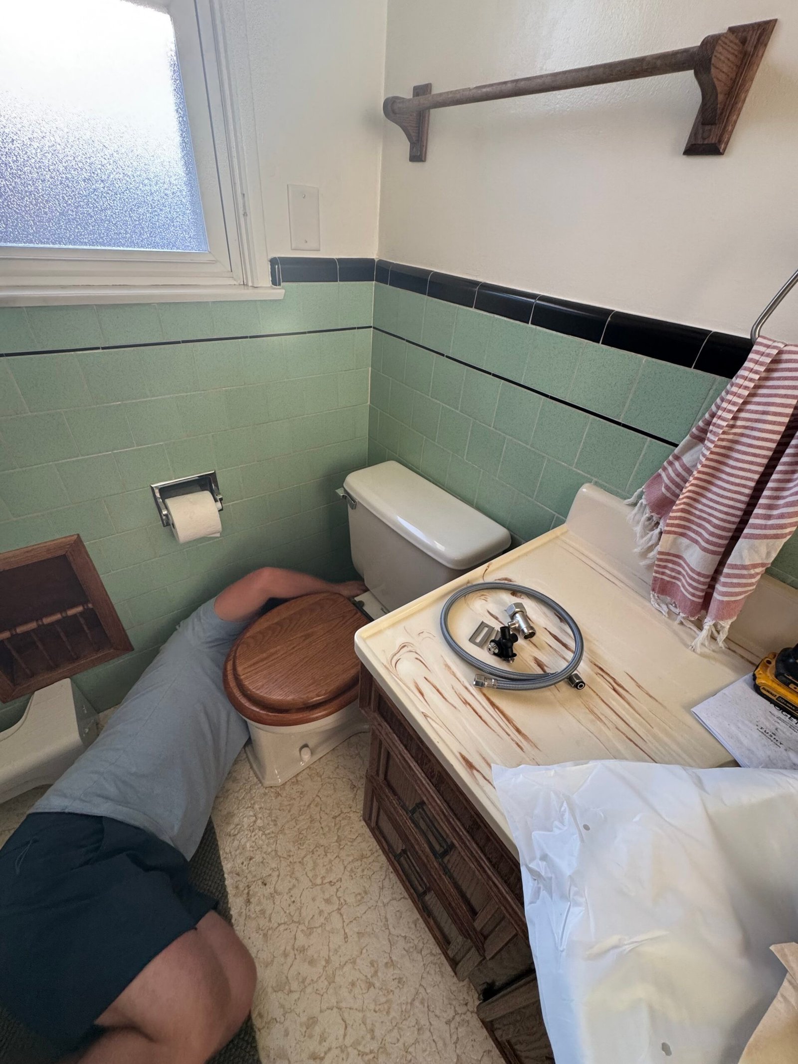 A person is lying on the bathroom floor next to a toilet, appearing to make repairs. The toilet seat is removed and placed on the sink, along with some tools.