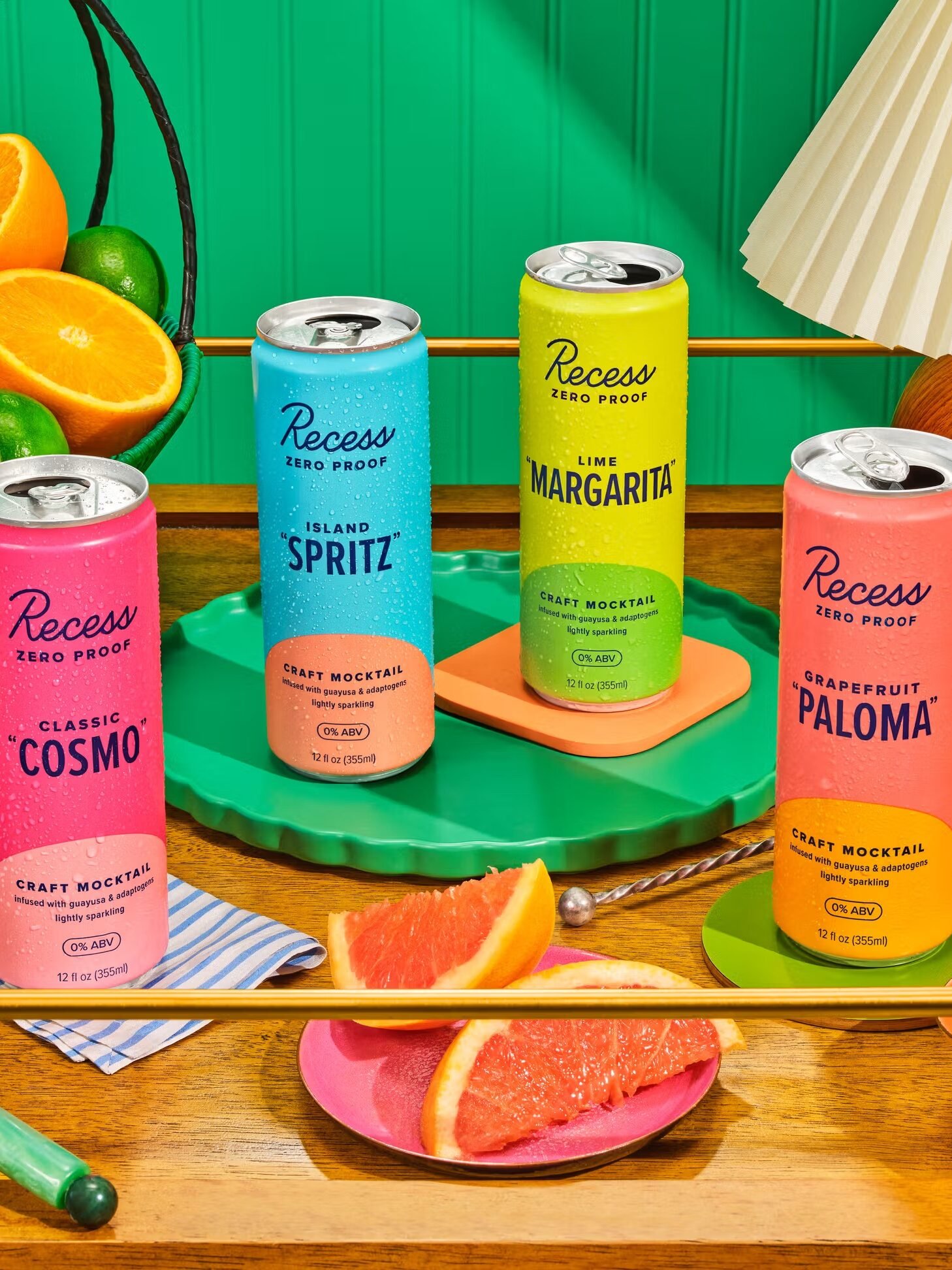 Four cans of Recess Zero Proof craft cocktails—Classic Cosmo, Island Spritz, Lime Margarita, and Grapefruit Paloma—are displayed on a wooden table with fresh fruit.