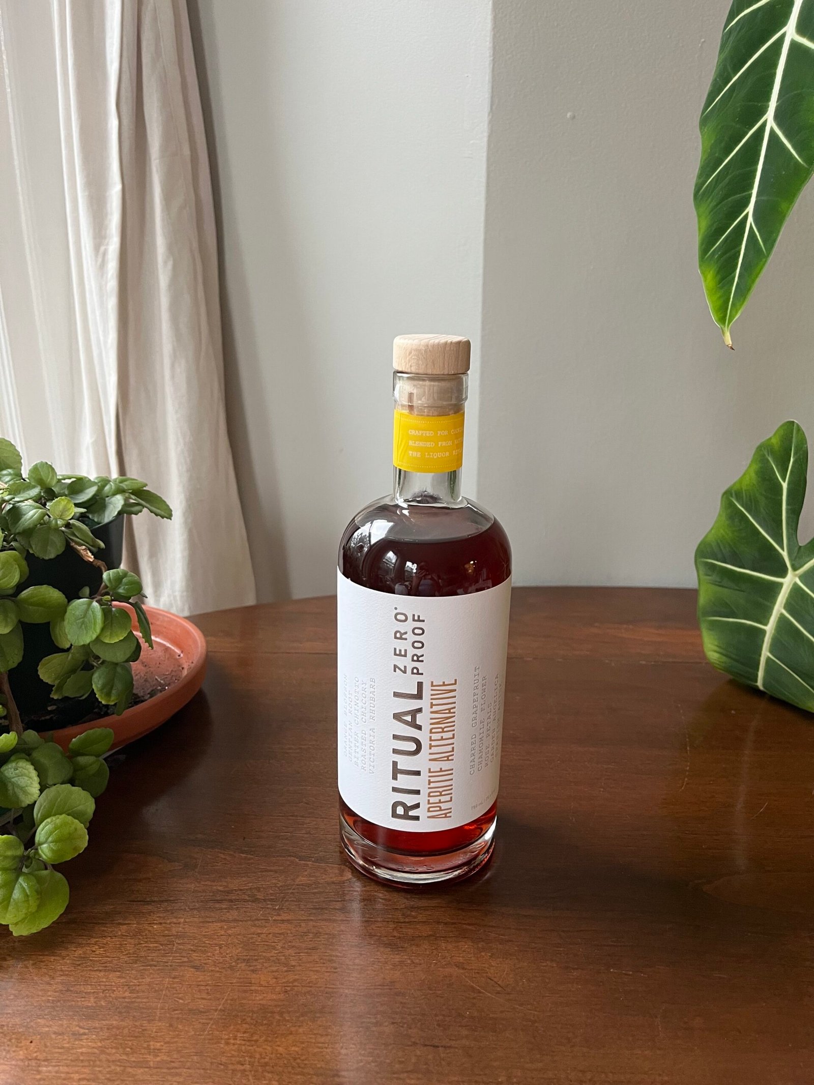 A glass bottle of Ritual Zero Proof whiskey alternative stands on a wooden surface. The label is white with black text. Green plants are in the background.