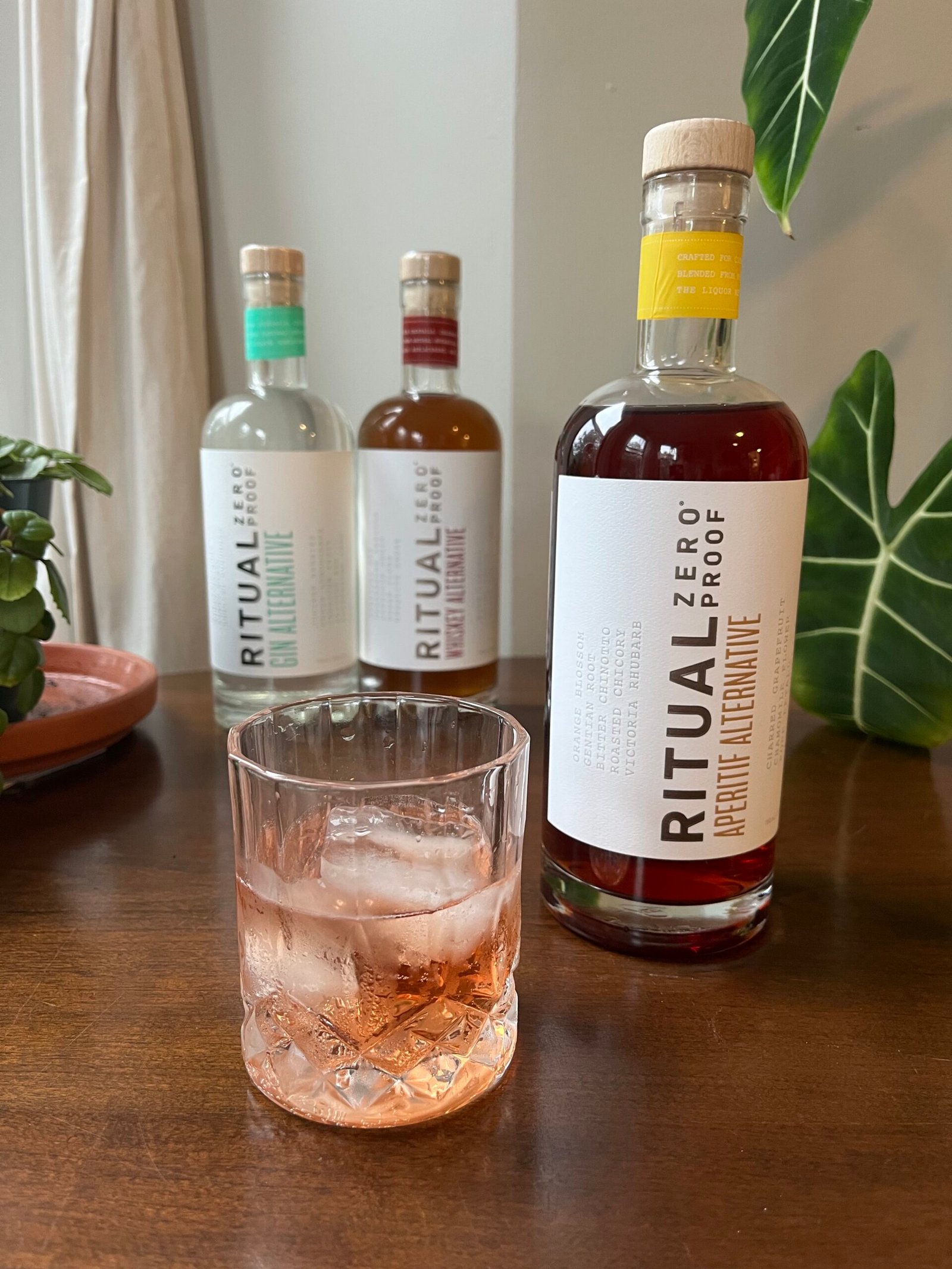 Cold beverage in a glass, with three bottles of Ritual Zero Proof beverages in the background on a wooden surface, next to a potted plant and large green leaf.