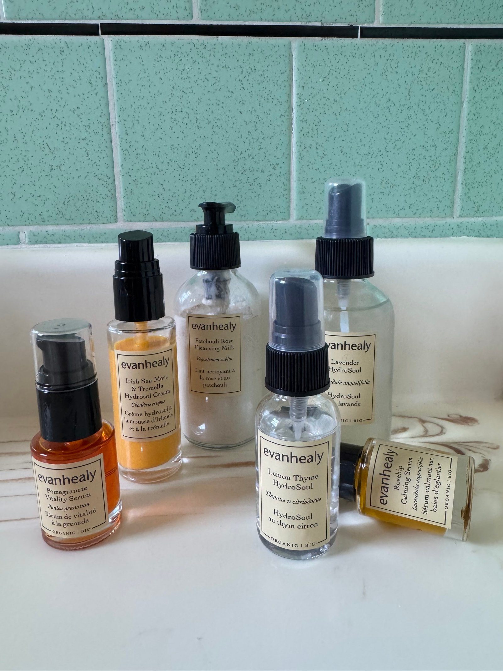 Six Evanhealy skincare bottles, including serums, cleansing milk, and hydrosols, arranged on a bathroom counter.