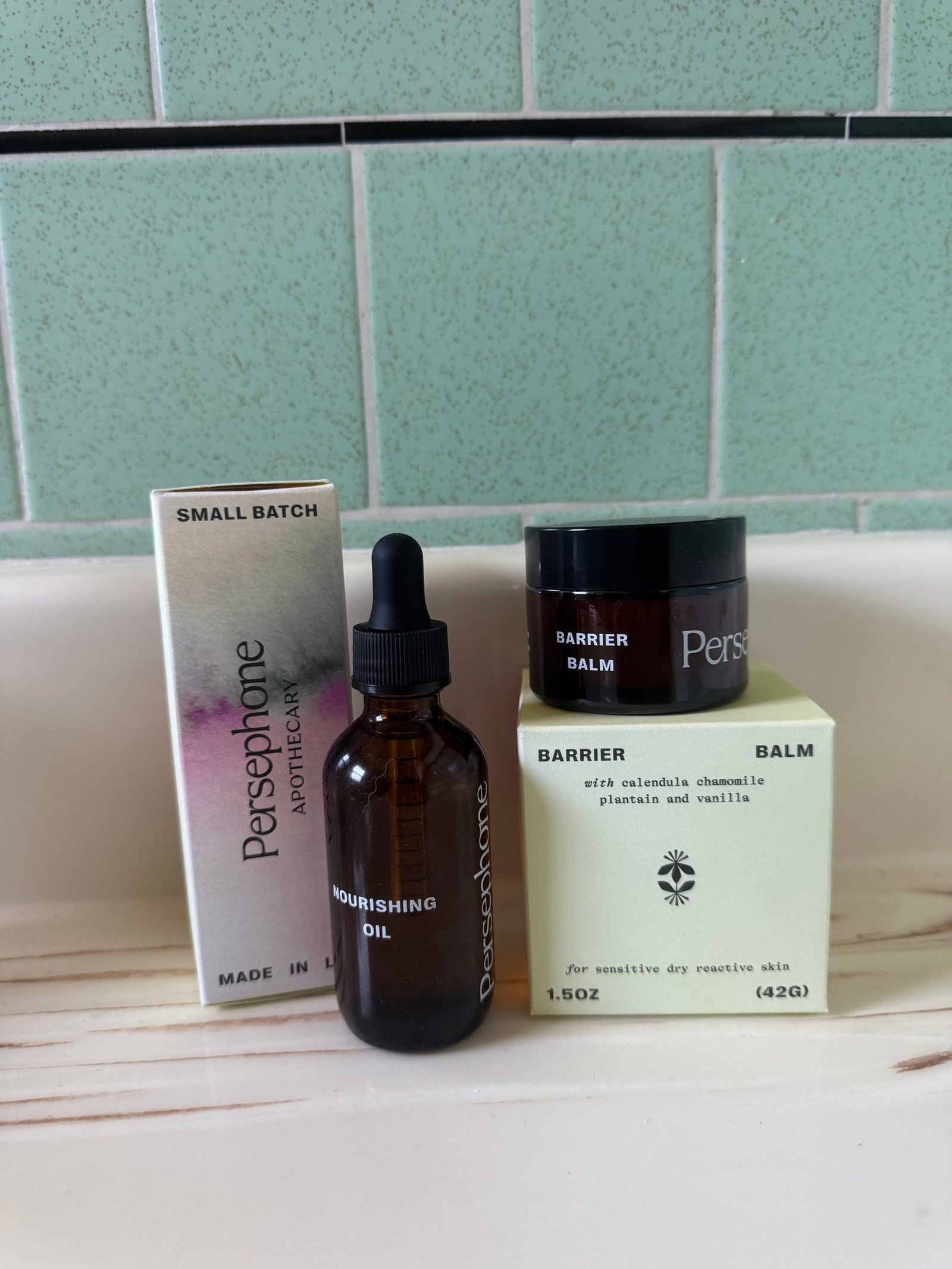 Persephone Apothecary Nourishing Oil in an amber dropper bottle with its box, and Barrier Balm in a jar with its box, sitting on a counter.