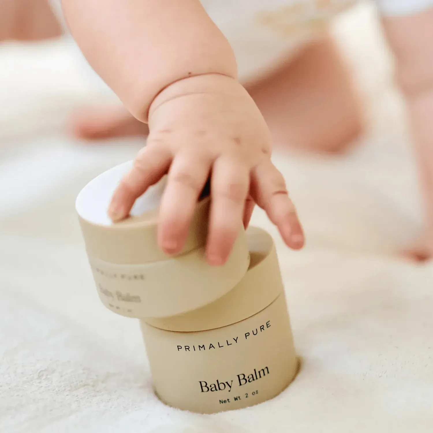A baby’s hand reaching for two stacked jars of Primally Pure Baby Balm.