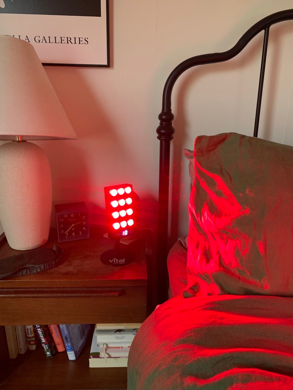 A red light therapy device is turned on and placed on a bedside table next to a lamp, clock, and bed with green bedding. Books are visible on a lower shelf.