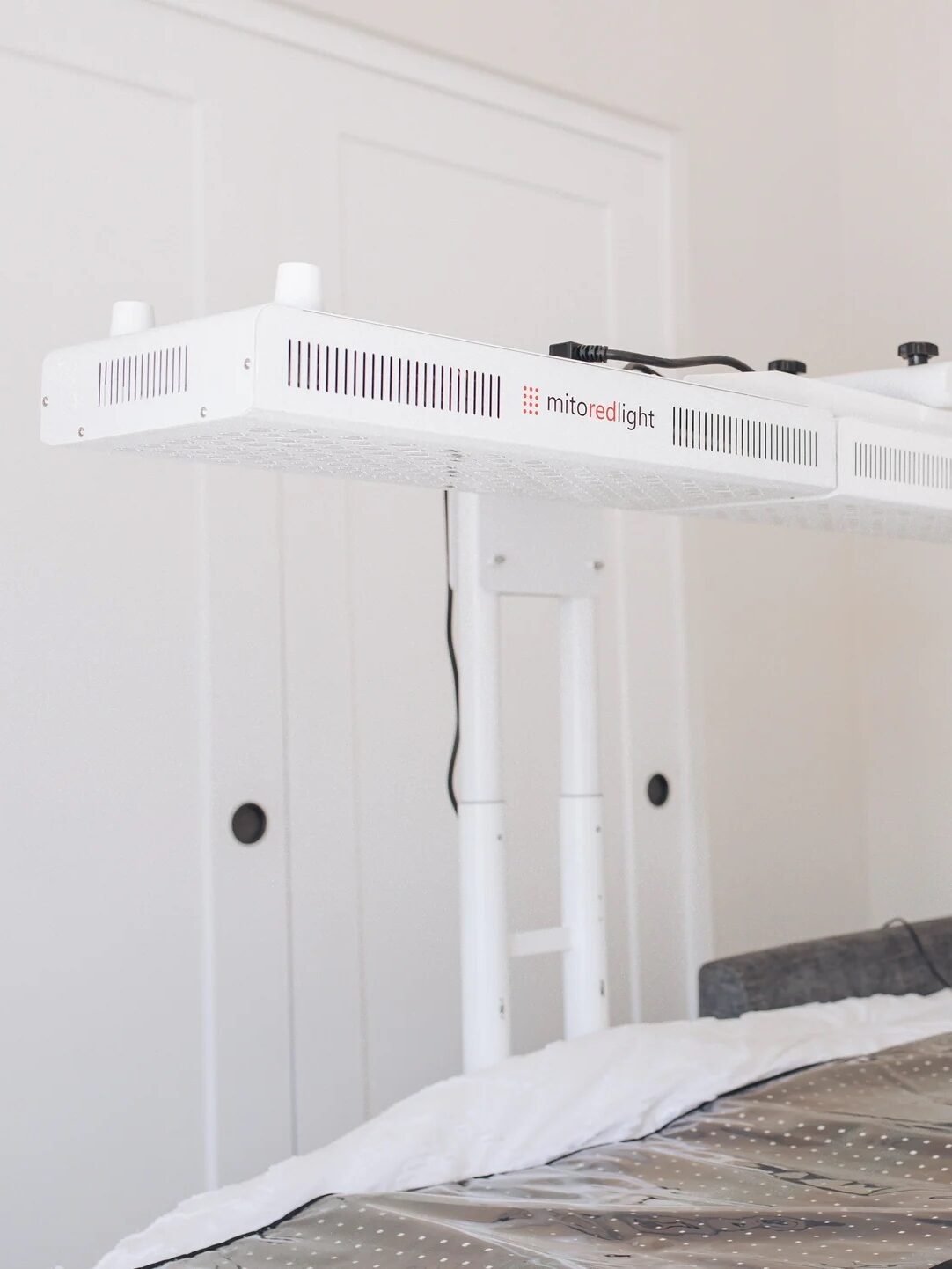 A bed with a white blanket and a wraparound blanket that says "REST. RELAX. REPEAT." has a large overhead LED light fixture suspended above it in a clean, white room.